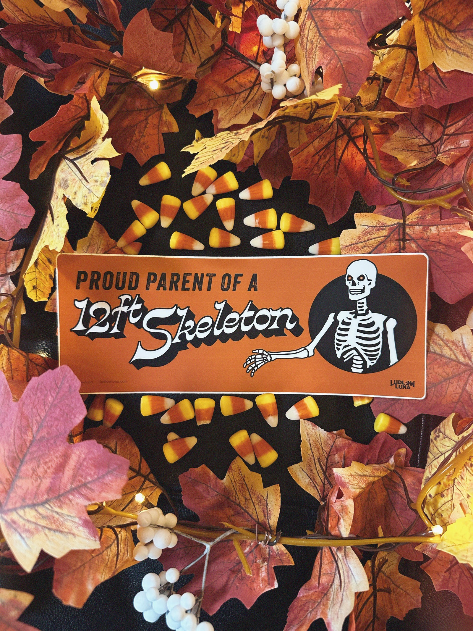Halloween-themed orange bumper sticker with a skeleton graphic that reads "Proud Parent of a 12ft Skeleton" on a background with candy corn and autumn leaves.