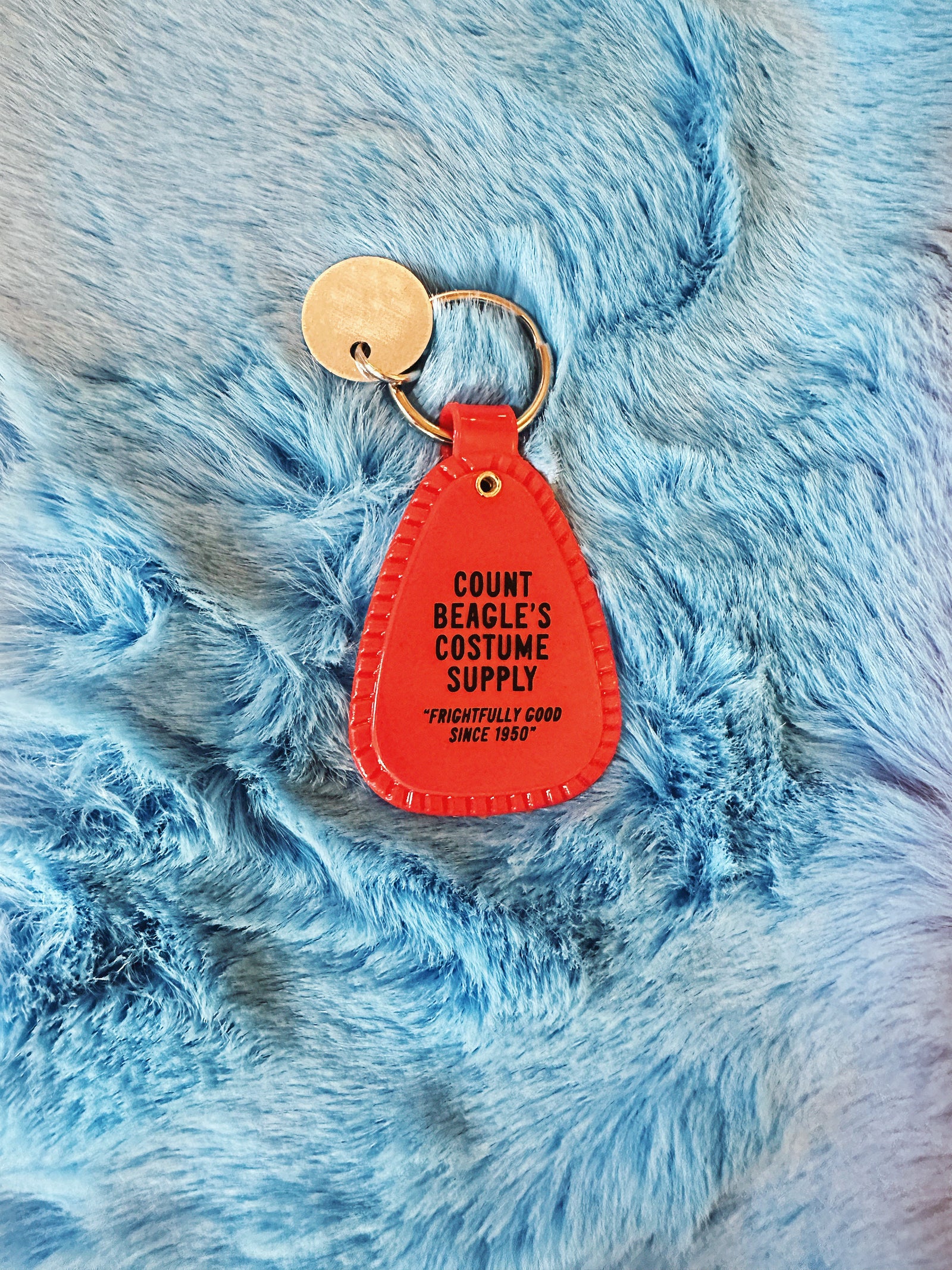 Black and Orange saddle style Halloween Snoopy Keychain reads "Count Beagle's Costume Supply. Frightfully good since 1950" on a fuzzy blue background.