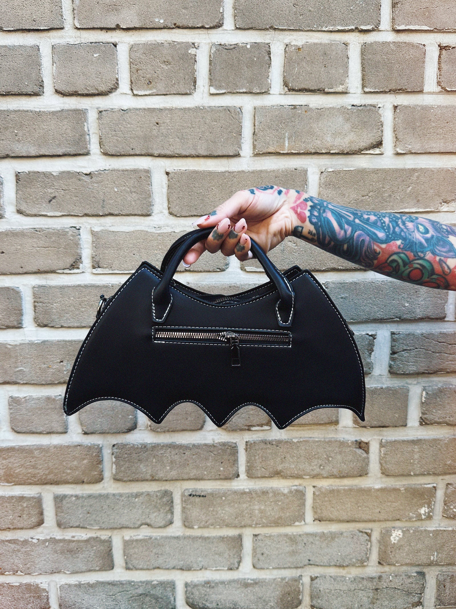 Black bat-shaped handbag held by a person with tattoos against a brick wall.