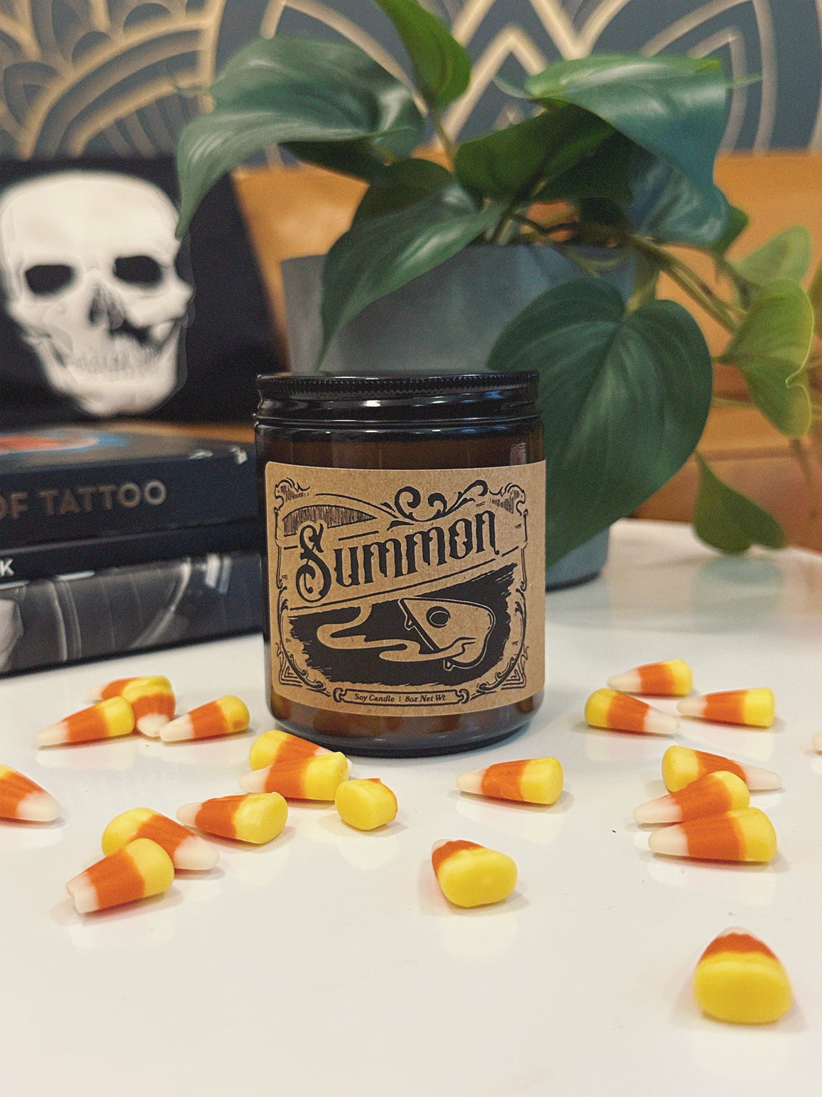 Jar candle labeled 'Summon' with planchette graphic label on a table with candy corn and plant in the background.