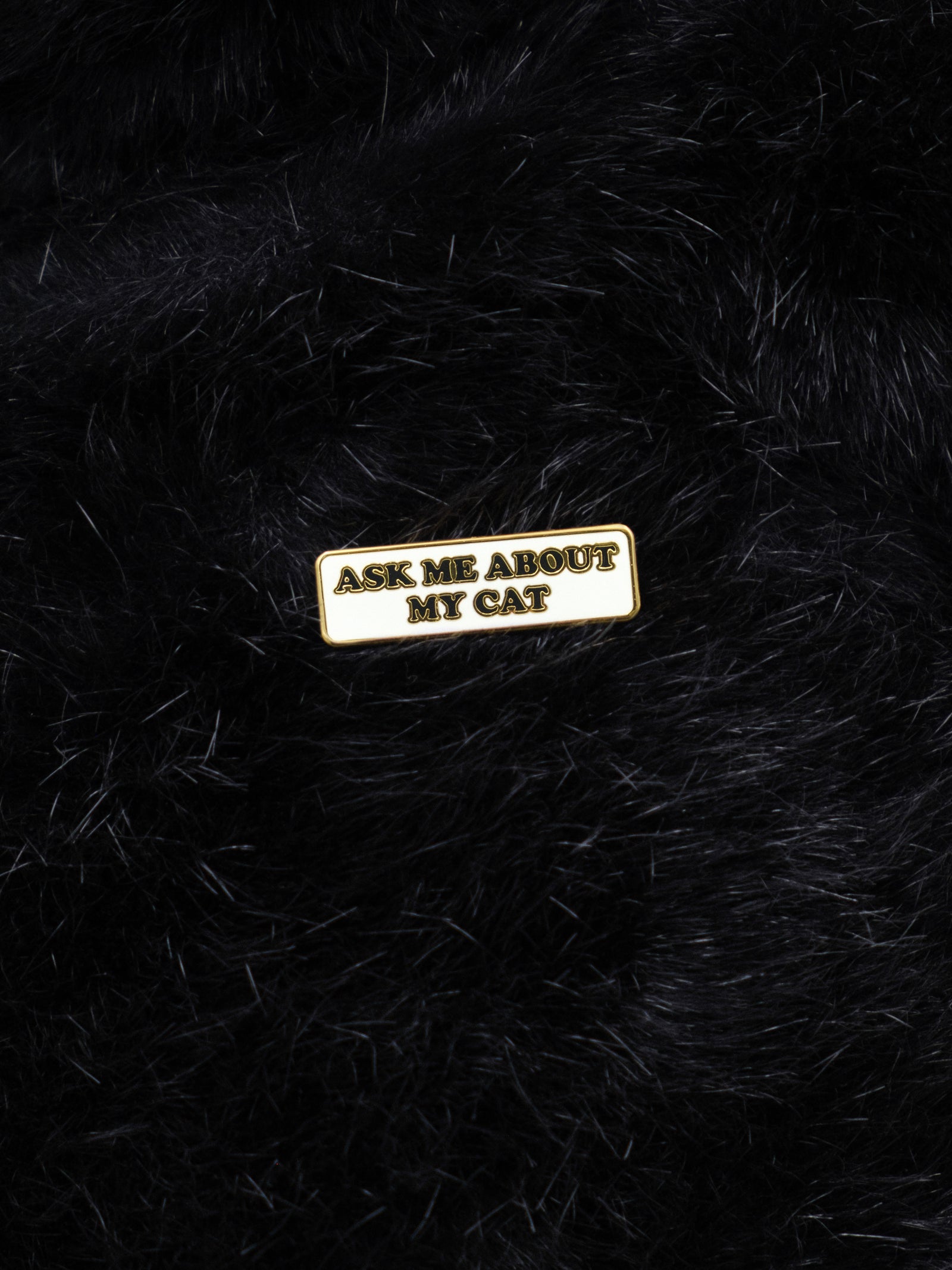 An enamel pin with the text 'ASK ME ABOUT MY CAT' attached to a black fuzzy background.