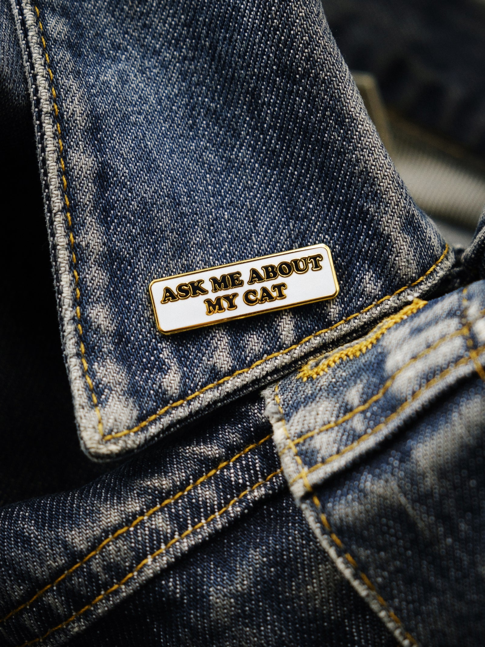 An enamel pin with the text 'ASK ME ABOUT MY CAT' attached to a denim fabric.