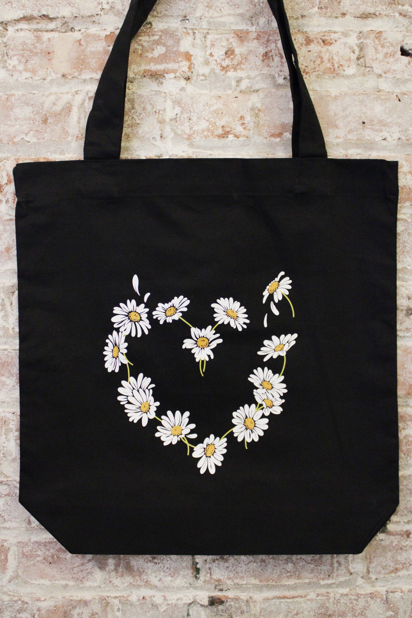 black cotton tote bag with daisy heart shaped graphic photographed against a brick wall.