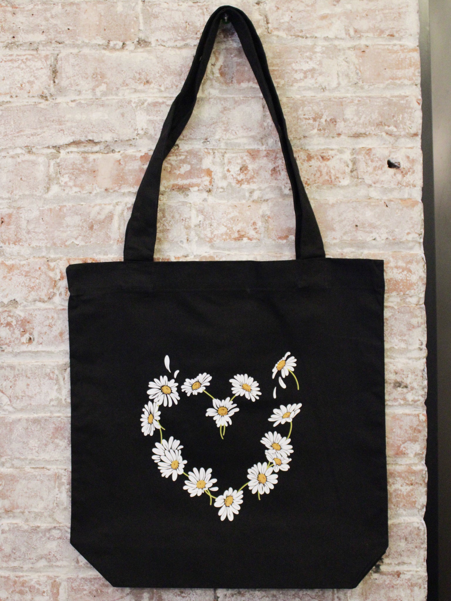 black cotton tote bag with daisy heart shaped graphic photographed against a brick wall.