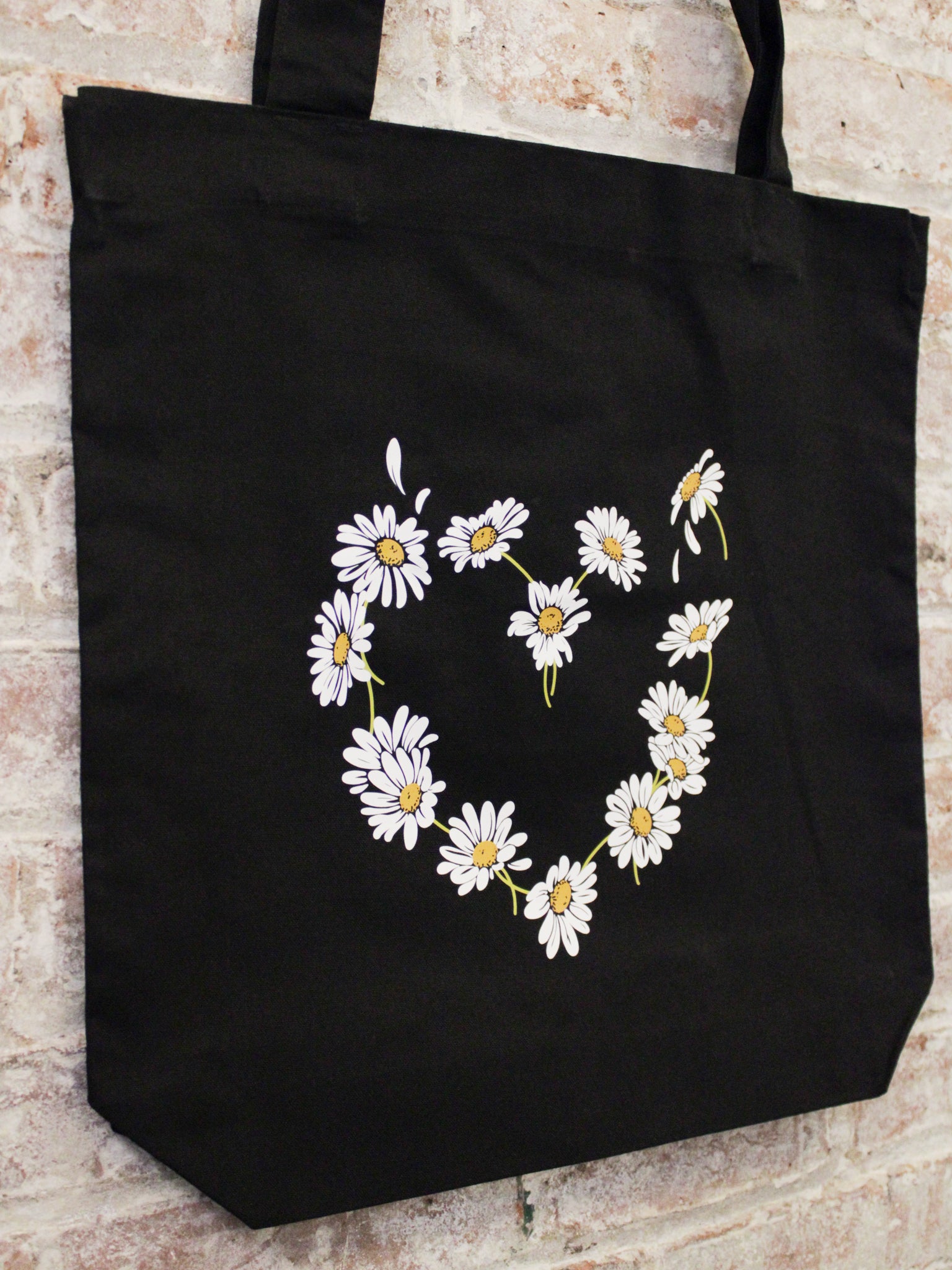 black cotton tote bag with daisy heart shaped graphic photographed at an angle against a brick wall.