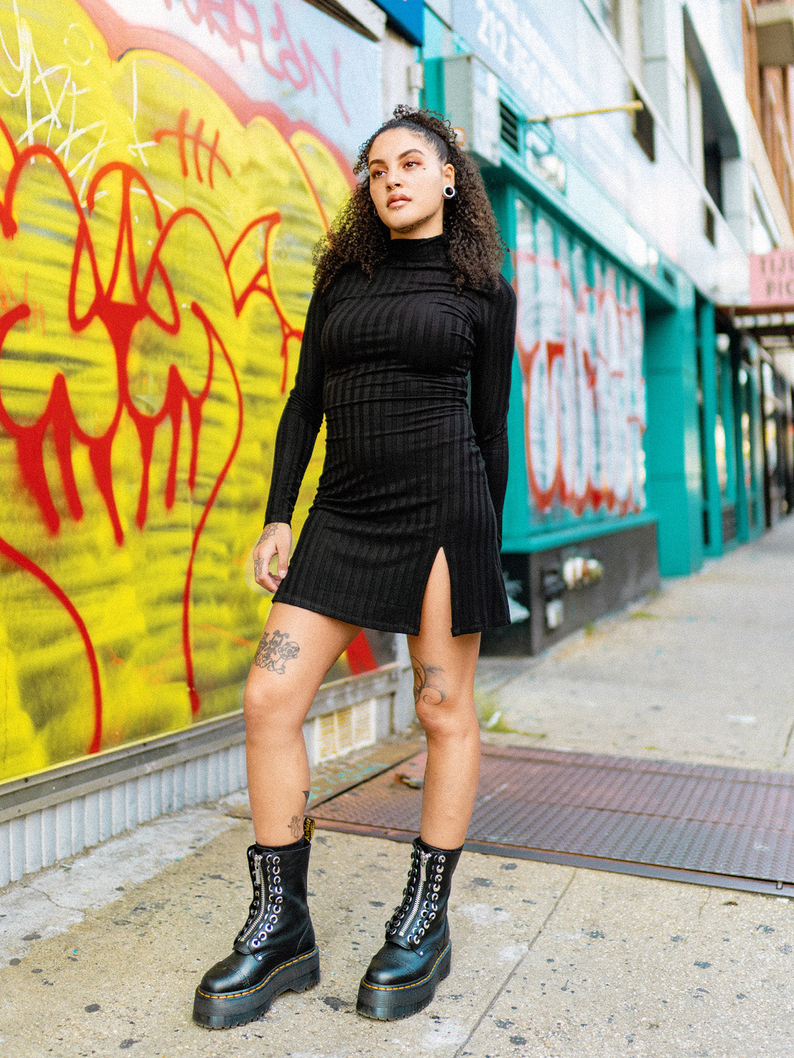 A person wearing a black mini dress with a mock neck, long sleeves, and a side slit. The dress has a bodycon fit and the person is paired with black boots, standing in front of a colorful graffiti wall.