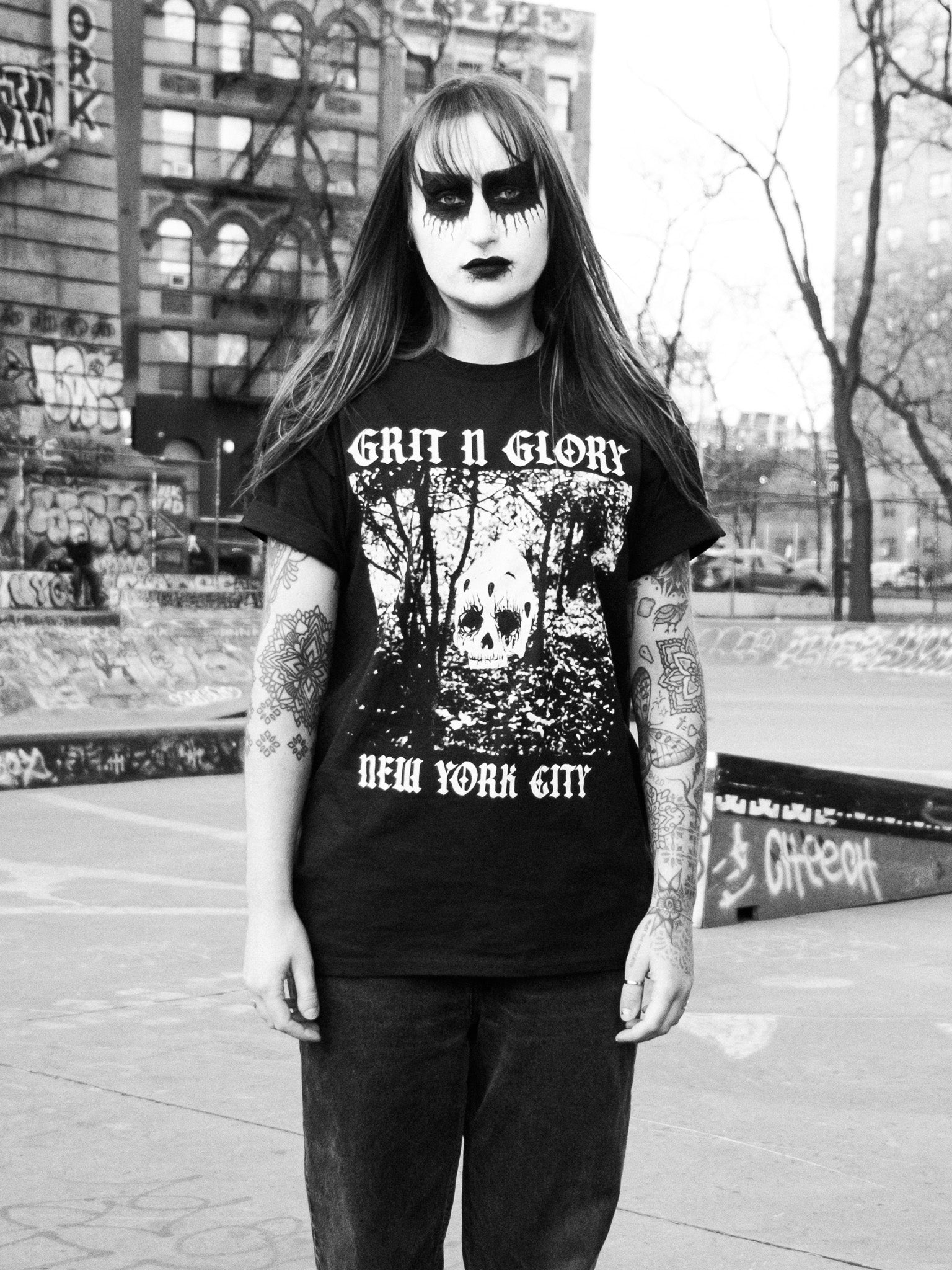 A person in black metal style makeup wearing an oversized black graphic tee with a skull and forest design and the text 'Grit N Glory New York City'.