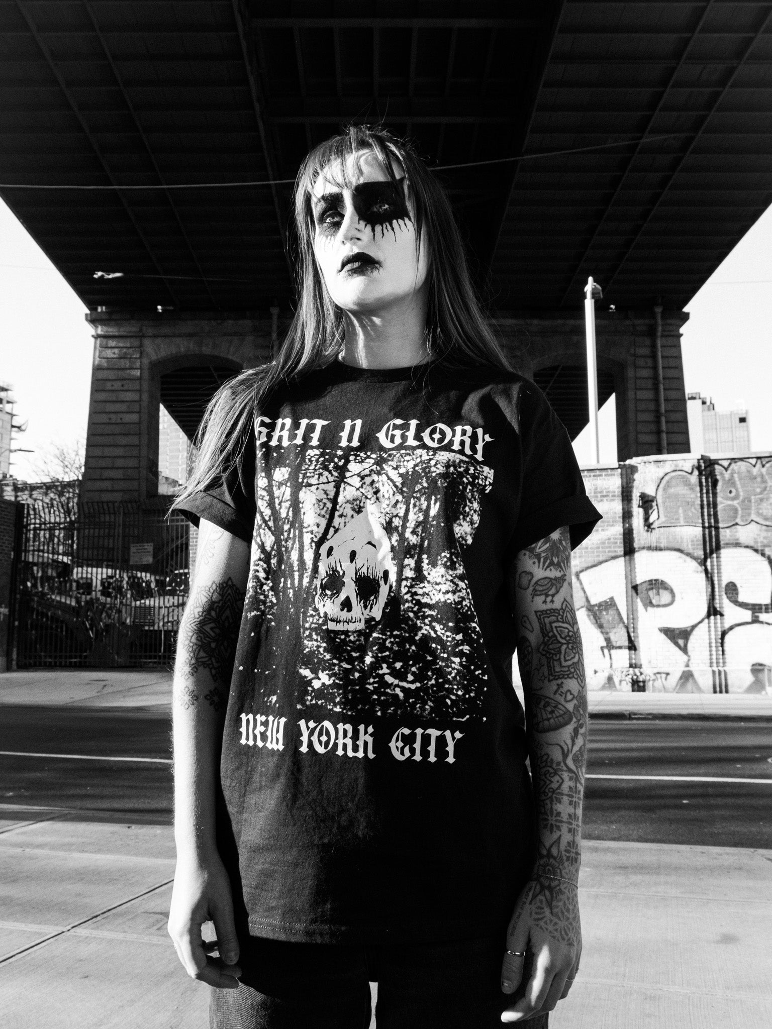 A person in black metal style makeup wearing an oversized black graphic tee with a skull and forest design and the text 'Grit N Glory New York City'.