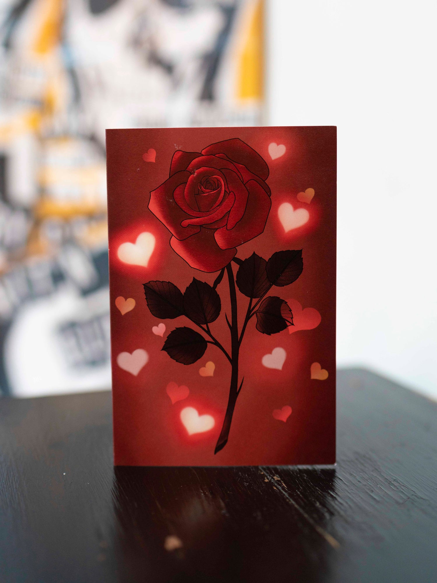 A folded greeting card with a red rose and hearts illustration on the front, with a pink envelope included.