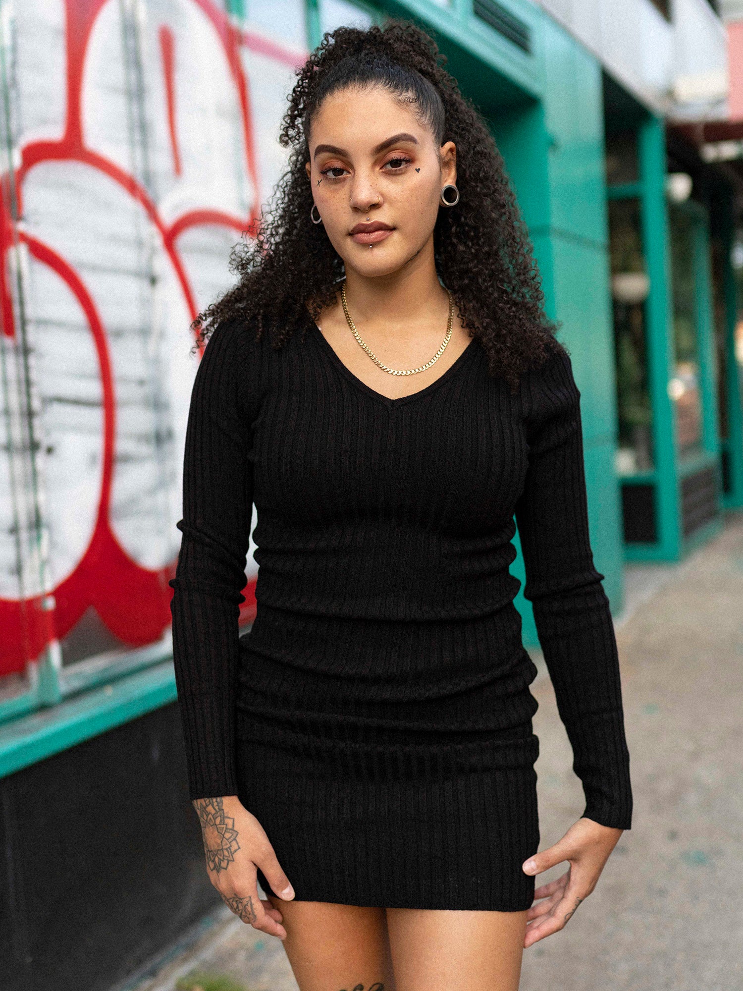 A woman standing on a sidewalk wearing a black long sleeve mini dress with a double v-neck, back neckline, and long sleeves. She is also wearing black boots and has tattoos visible on her arms.