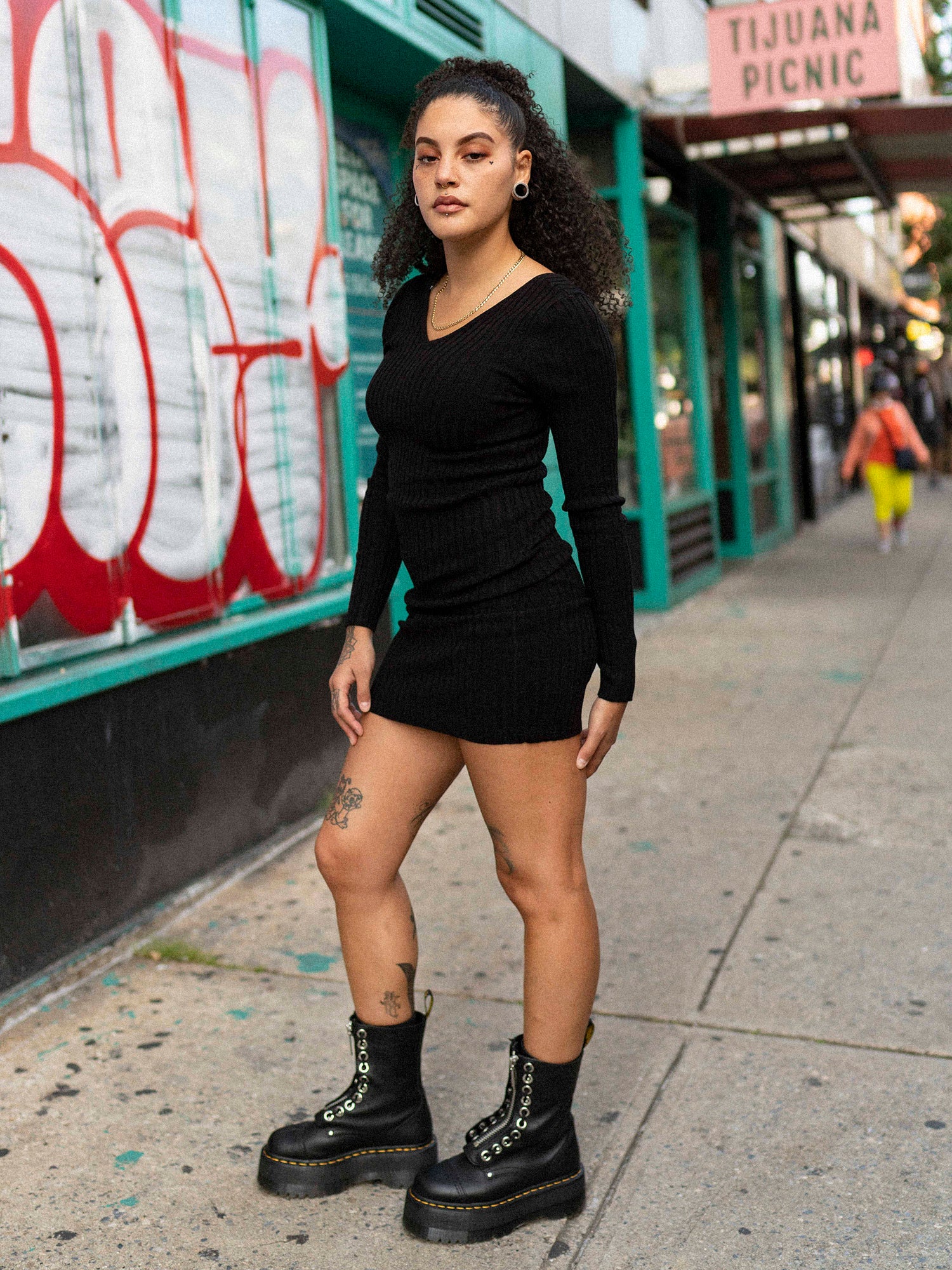 A woman standing on a sidewalk wearing a black long sleeve mini dress with a double v-neck, back neckline, and long sleeves. She is also wearing black boots and has tattoos visible on her arms.