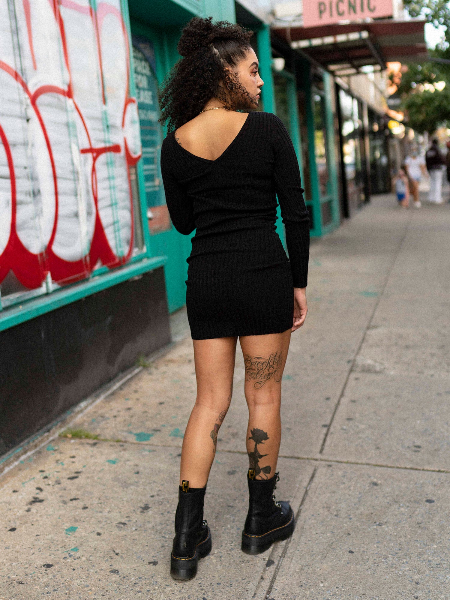 A woman standing on a sidewalk wearing a black long sleeve mini dress with a double v-neck, back neckline, and long sleeves. She is also wearing black boots and has tattoos visible on her arms.