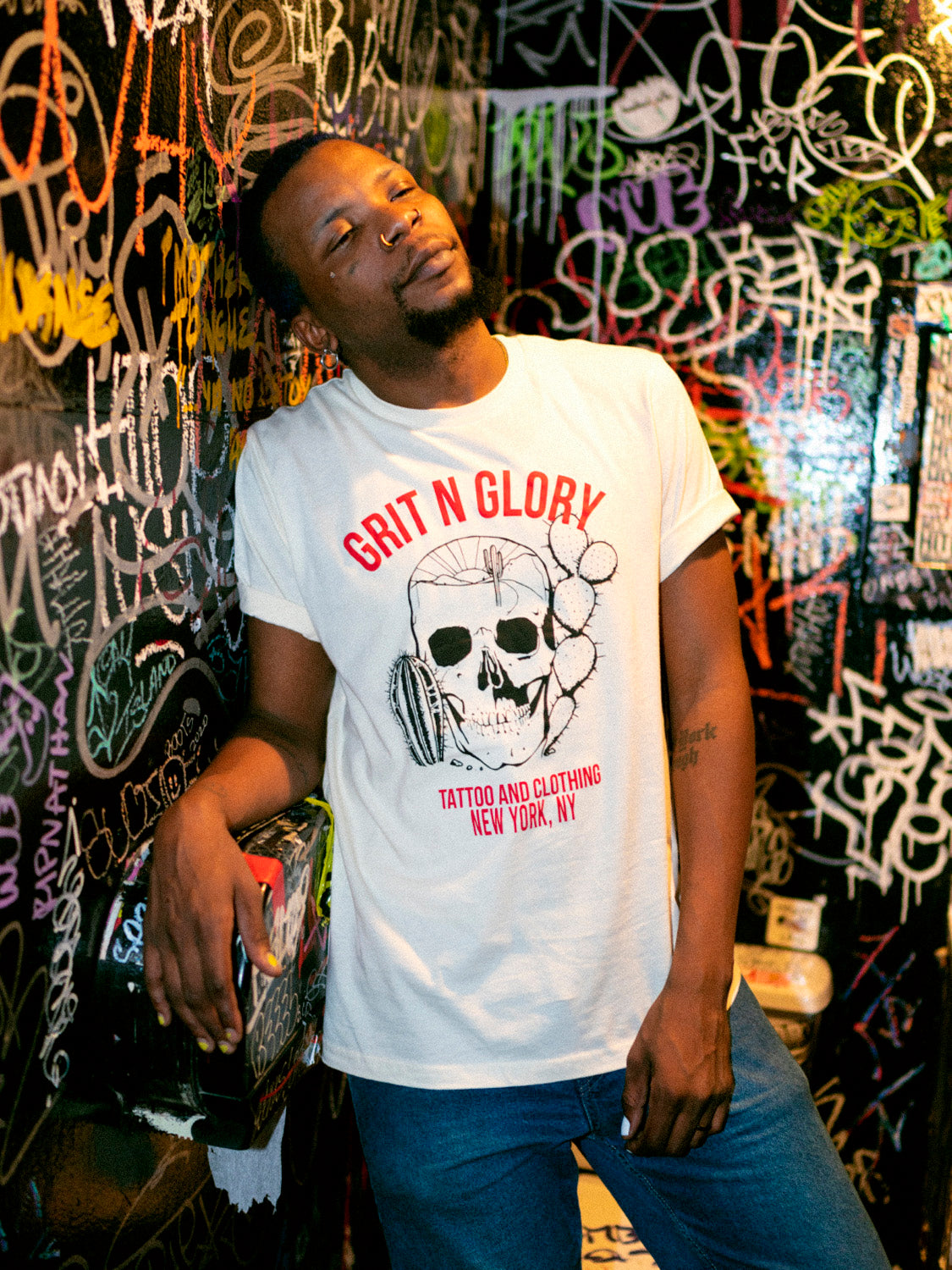 A person wearing a white crew neck t-shirt with a graphic design of a skull and text, standing in front of a graffiti wall.