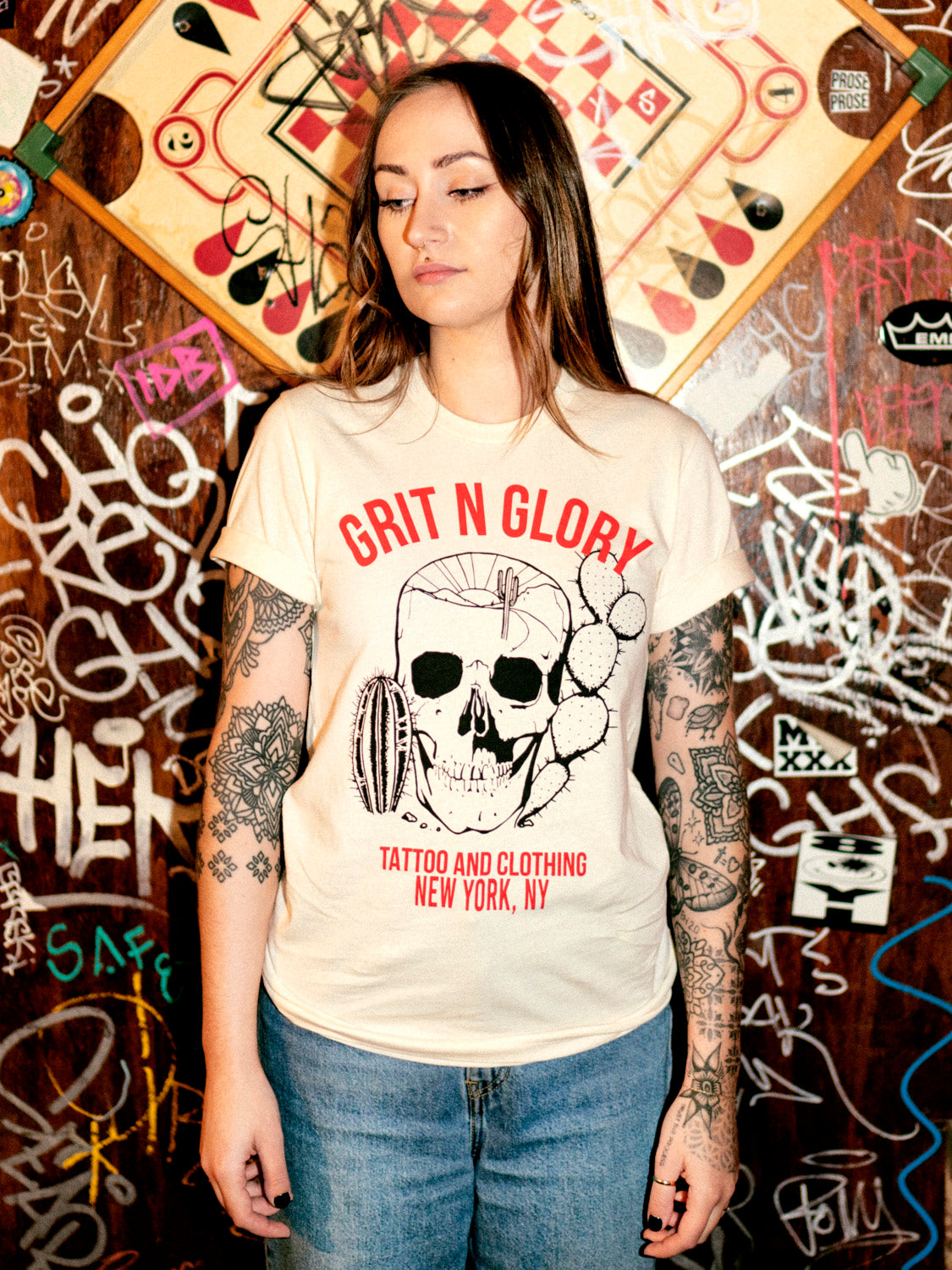 A person wearing a white crew neck t-shirt with a graphic design of a skull, cacti and Grit N Glory red text in front of a dive bar graffiti wall.
