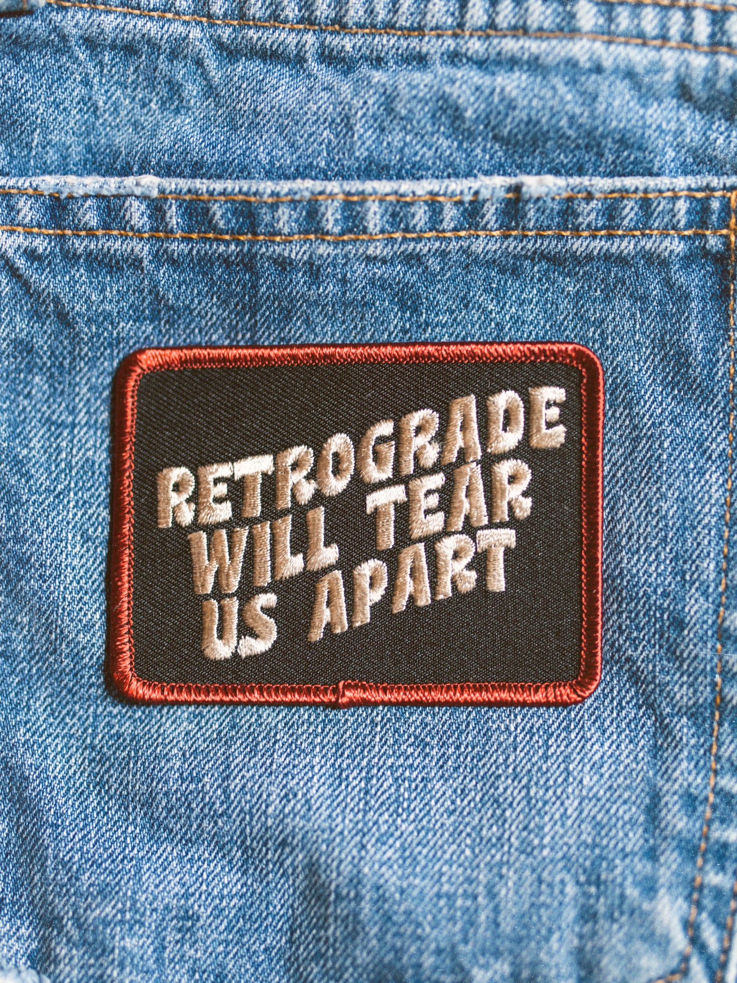 A canvas embroidered patch with text 'RETROGRADE WILL TEAR US APART' ironed onto a denim fabric.