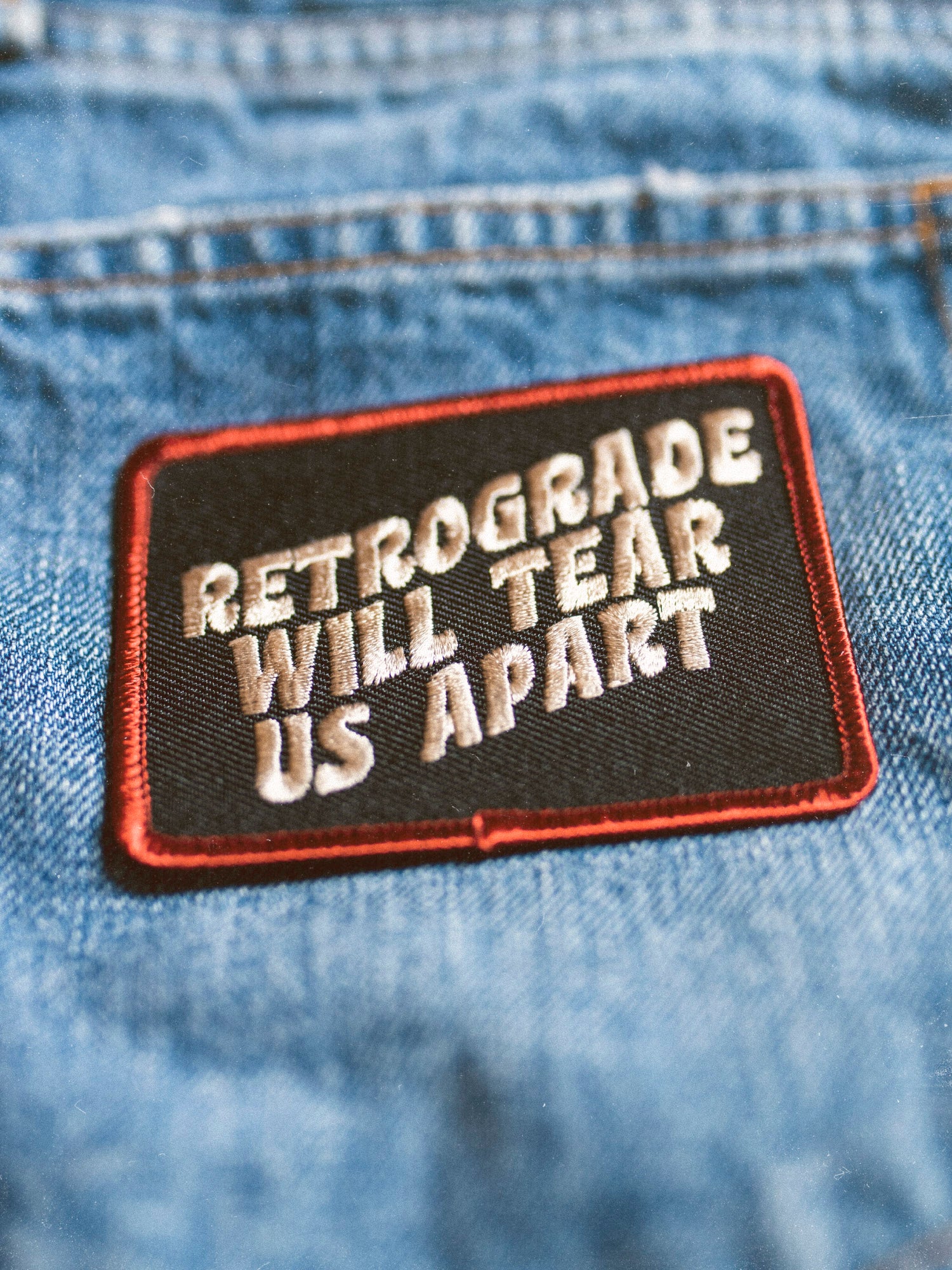 A canvas embroidered patch with text 'RETROGRADE WILL TEAR US APART' ironed onto a denim fabric.