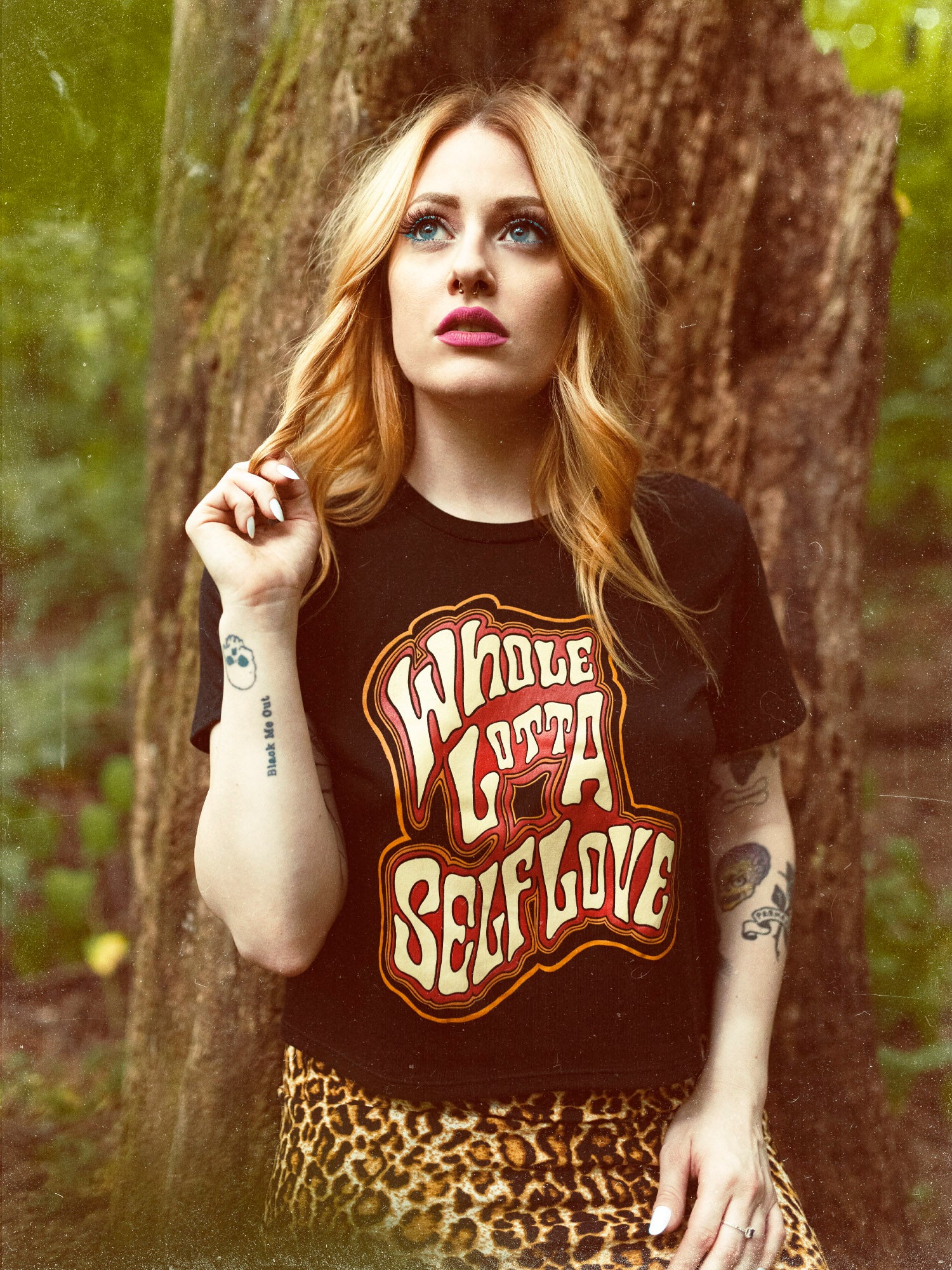 A female model wearing a black crop top with the graphic 'Whole Lotta Self Love' standing next to a tree.
