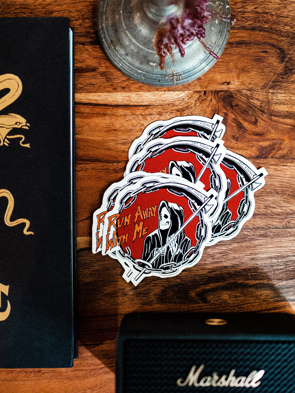 A die cut vinyl sticker with a black and white reaper design in a red circle, placed on a wooden surface next to a Marshall speaker.
