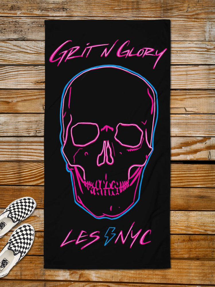 An oversized beach towel with a neon skull graphic in black, pink, and blue on a wooden floor, with the brand 'Grit N Glory' and location 'LES NYC' printed below the skull.