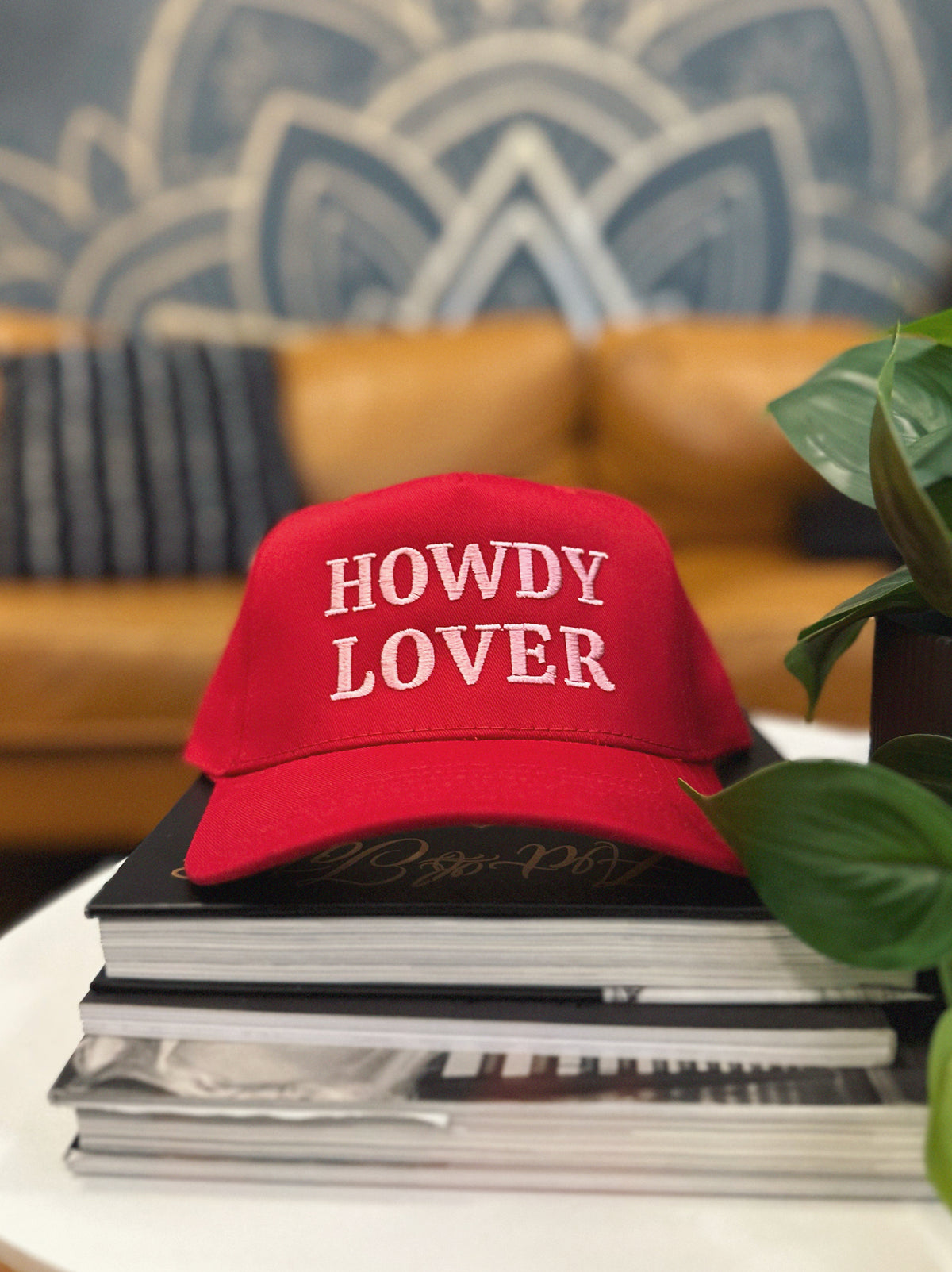 Red cap with 'HOWDY LOVER' pink text on a stack of books with a decorative wall in the background