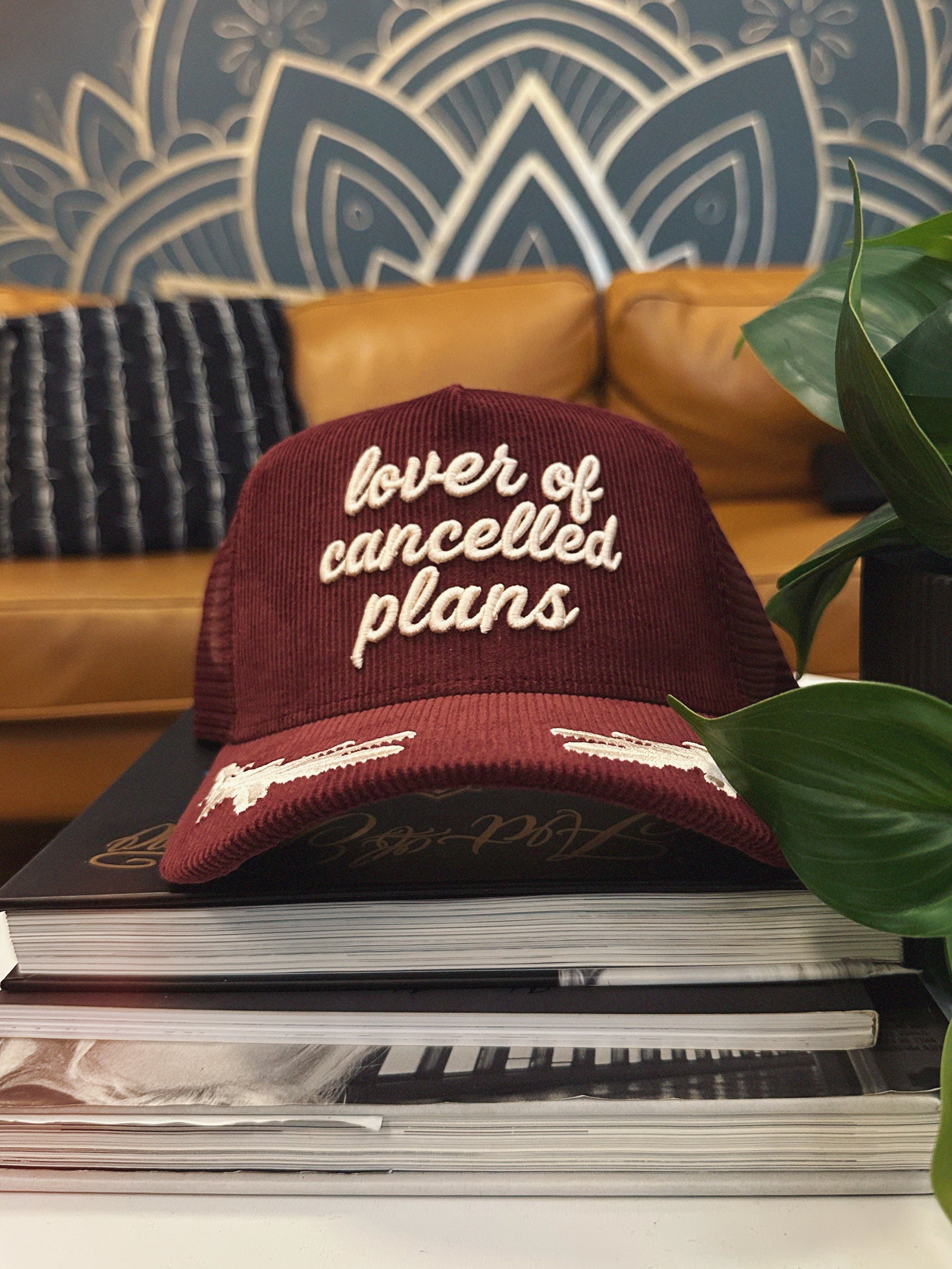 Maroon cap with 'lover of cancelled plans' embroidered text on a stack of books against a decorative wall.