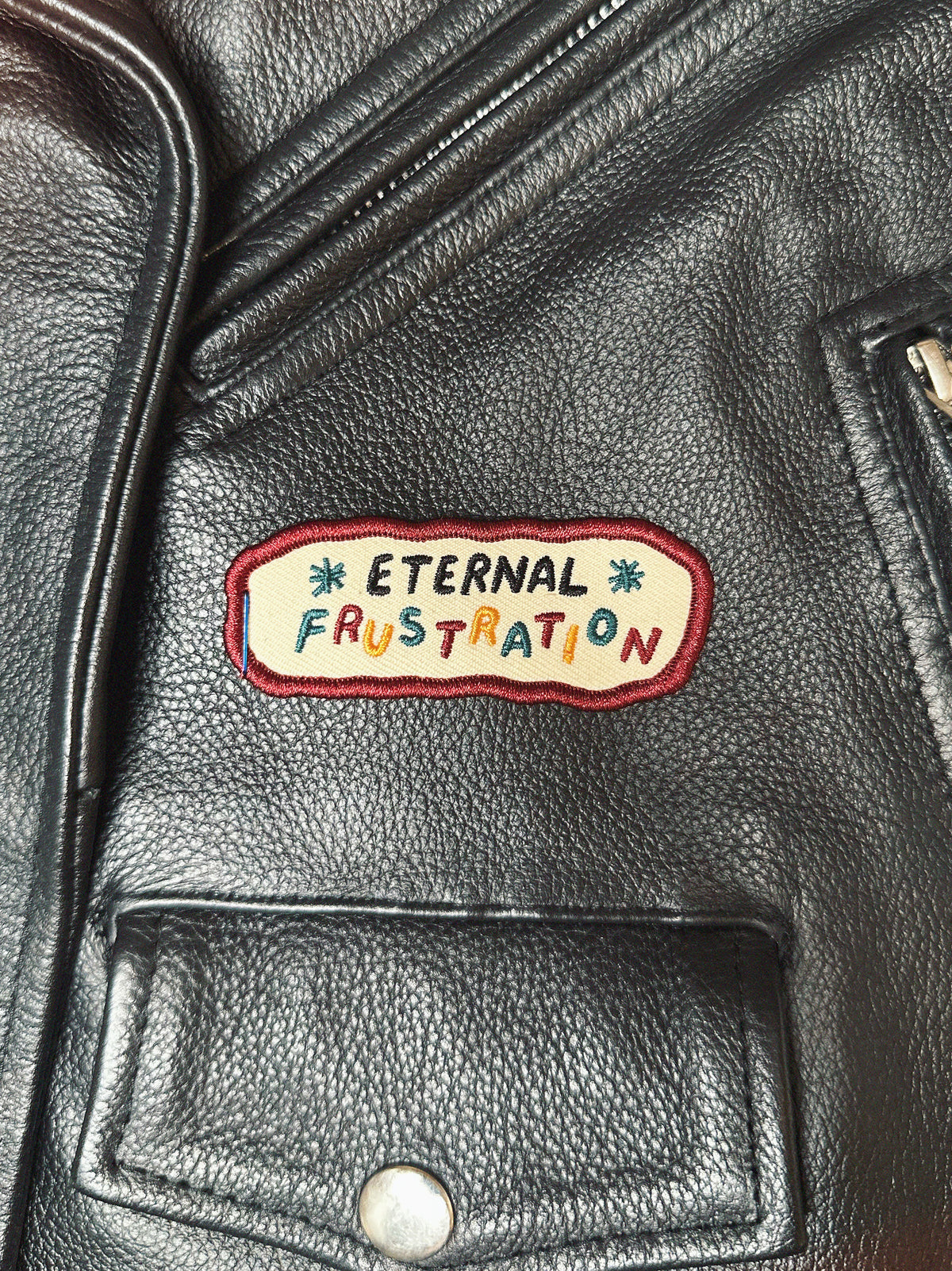 Black leather moto jacket with a colorful patch reading 'Eternal Frustration'.