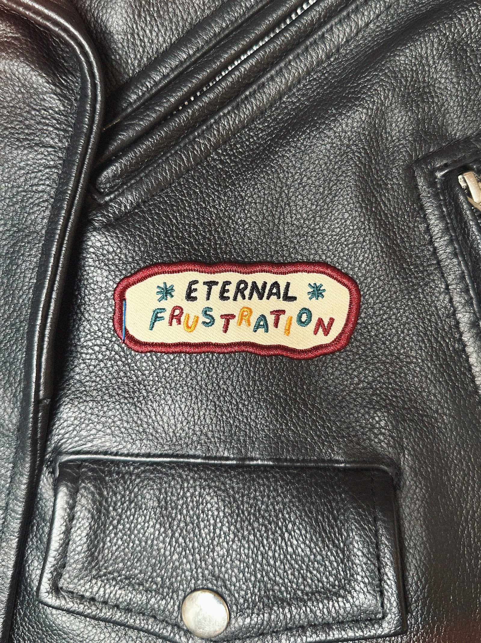 Black leather moto jacket with a colorful patch reading 'Eternal Frustration'.