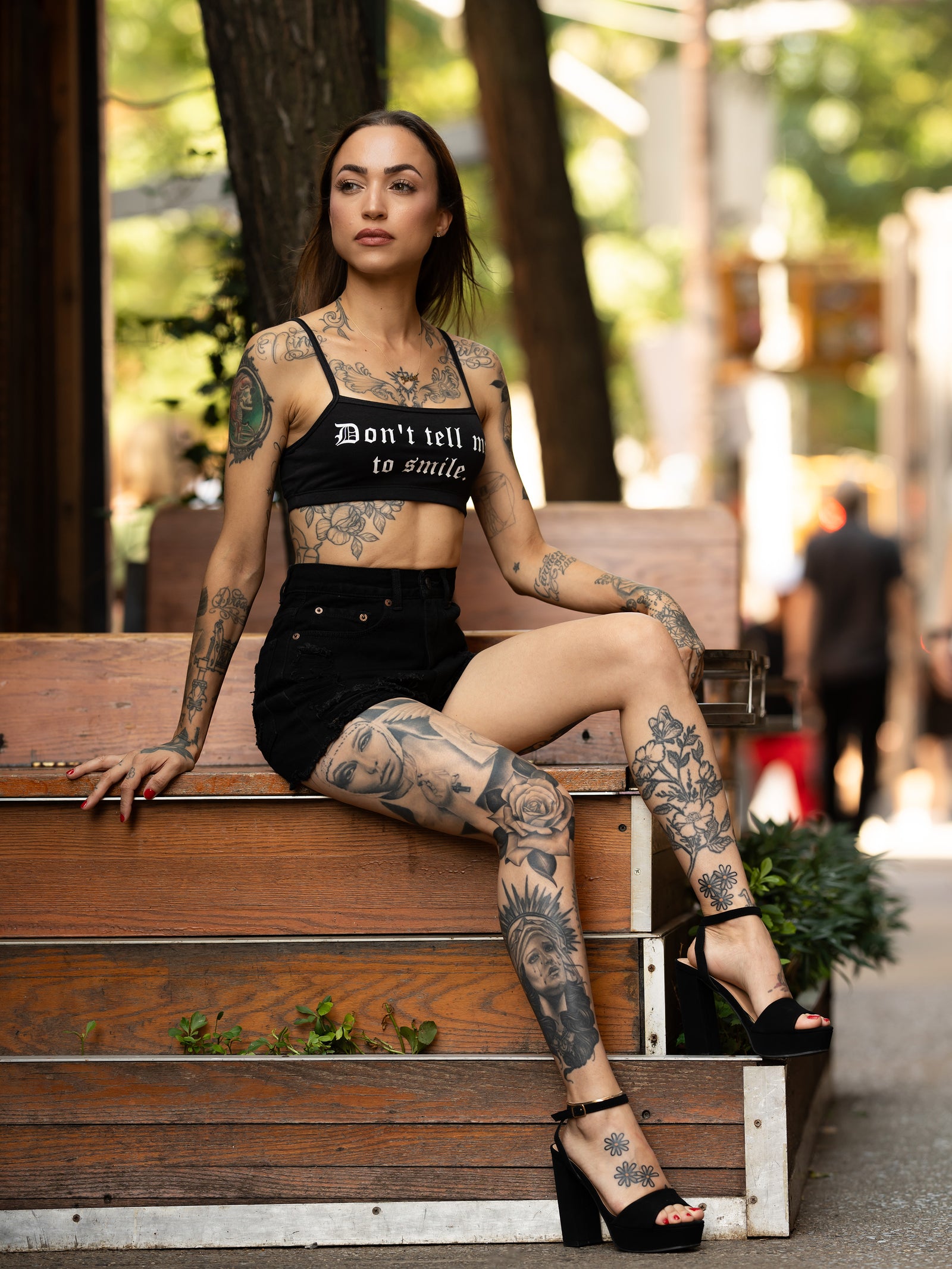 Janice Danger wearing a "Dont Tell Me To Smile" black bralette with black denim shorts, seated on a bench. 