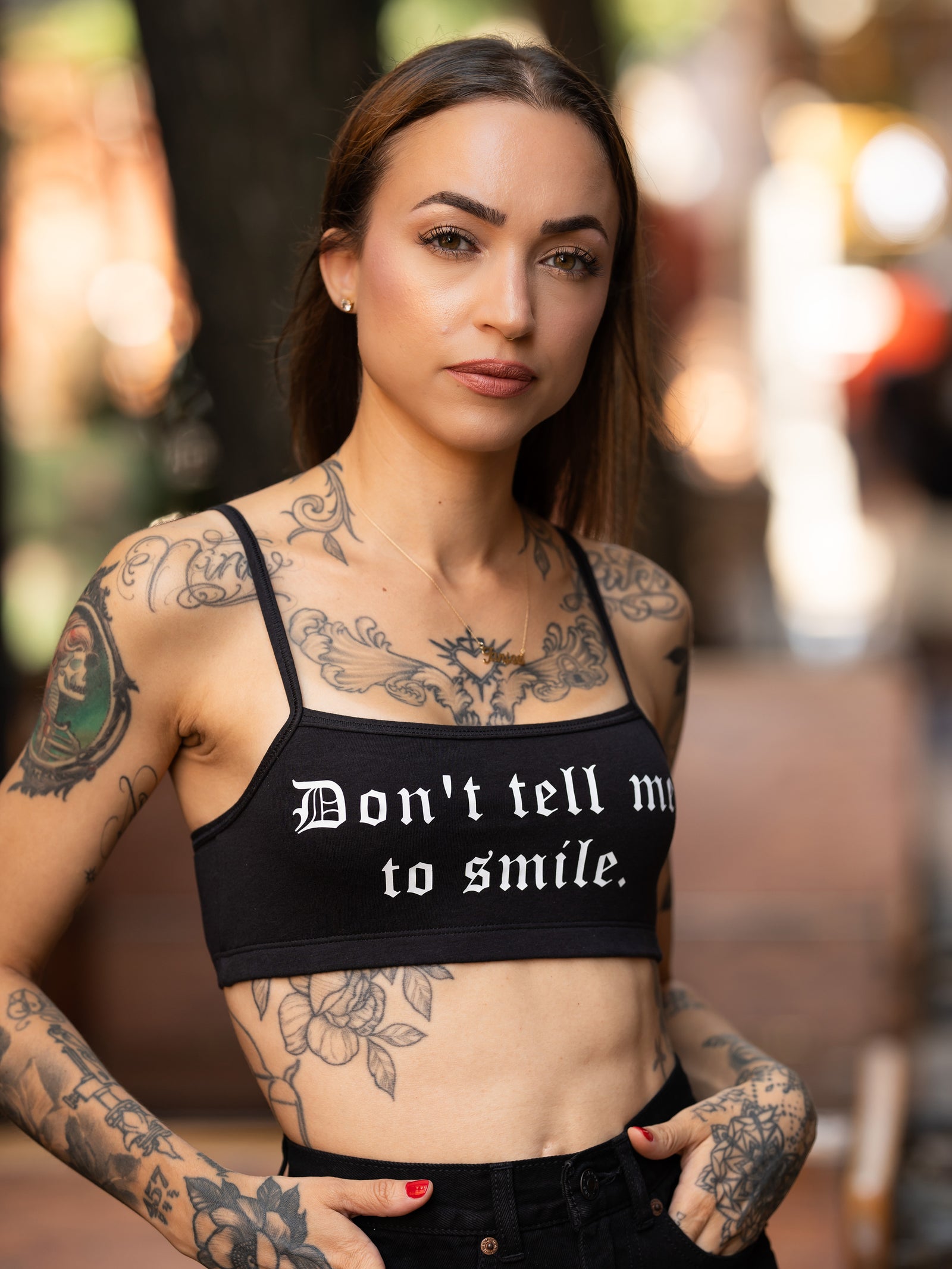 Janice Danger wearing a "Dont Tell Me To Smile" black bralette with black denim shorts, seated on a bench. 