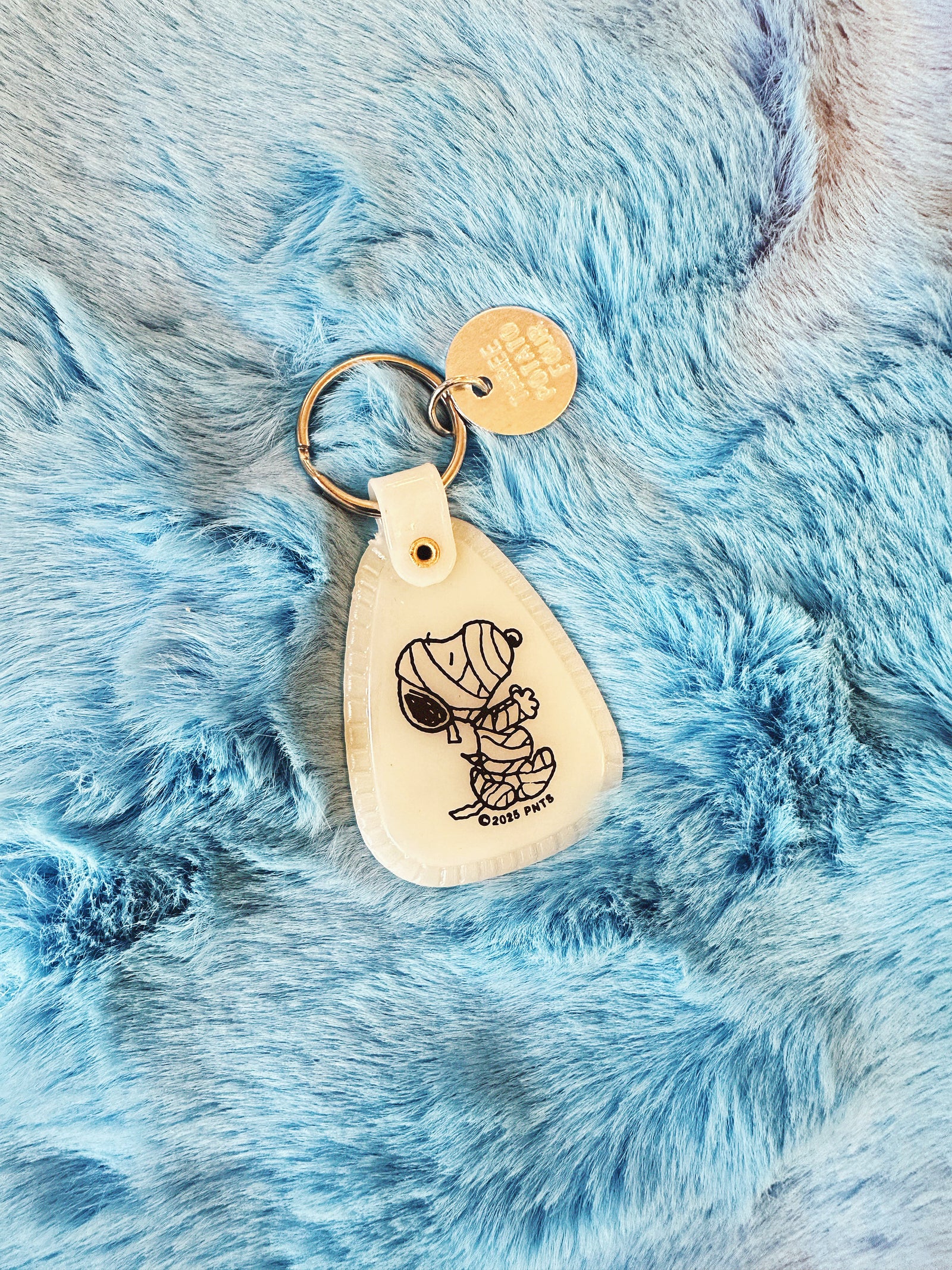 Saddle style, glow in the dark, Halloween Snoopy Mummy keychain on a fuzzy blue background. 