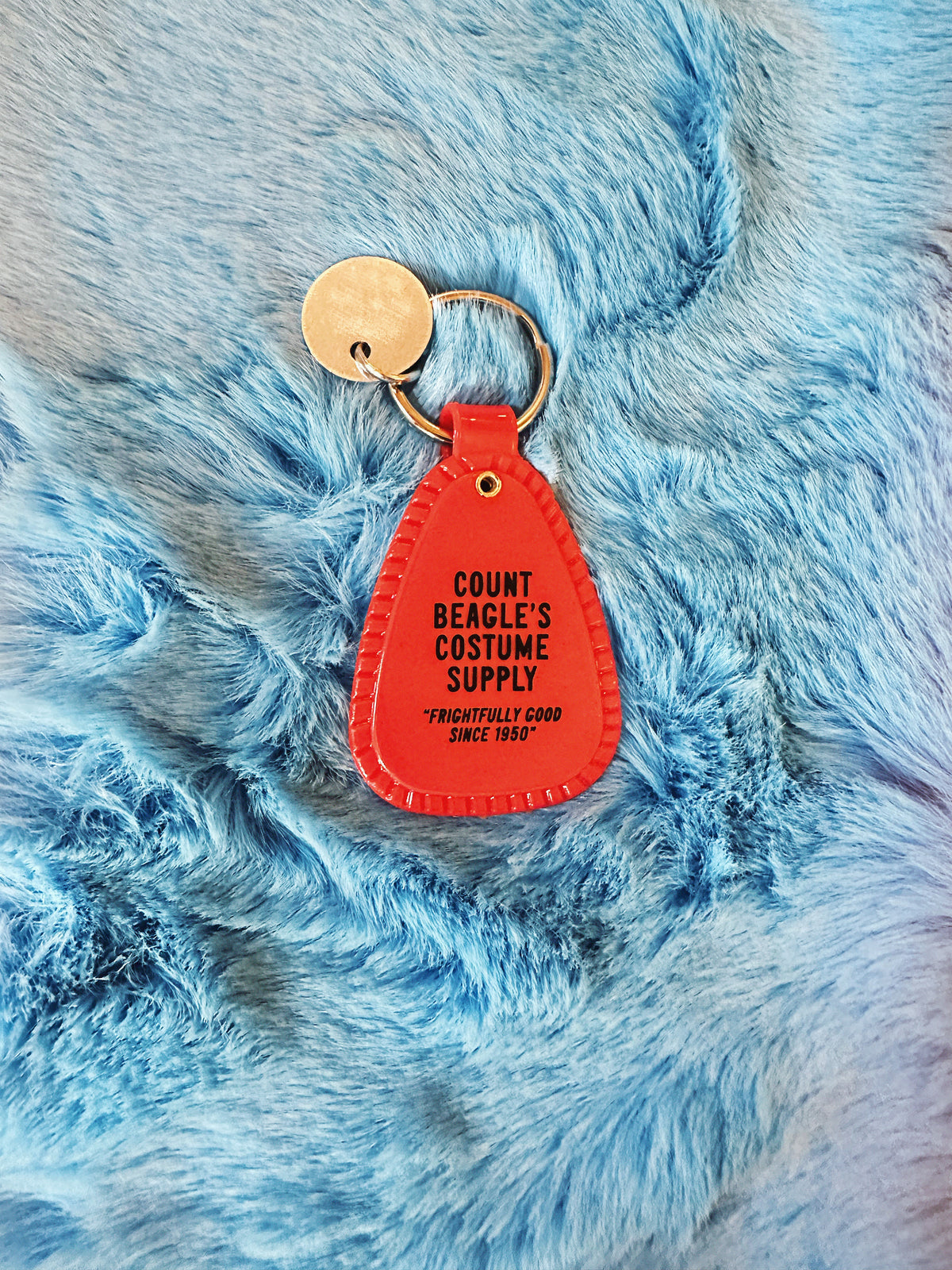 Black and Orange saddle style Halloween Snoopy Keychain reads "Count Beagle's Costume Supply. Frightfully good since 1950" on a fuzzy blue background.