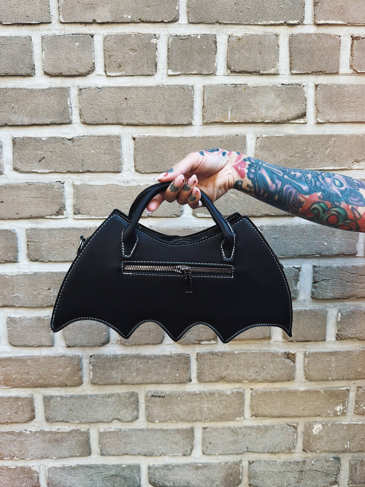 Black bat-shaped handbag held by a person with tattoos against a brick wall.