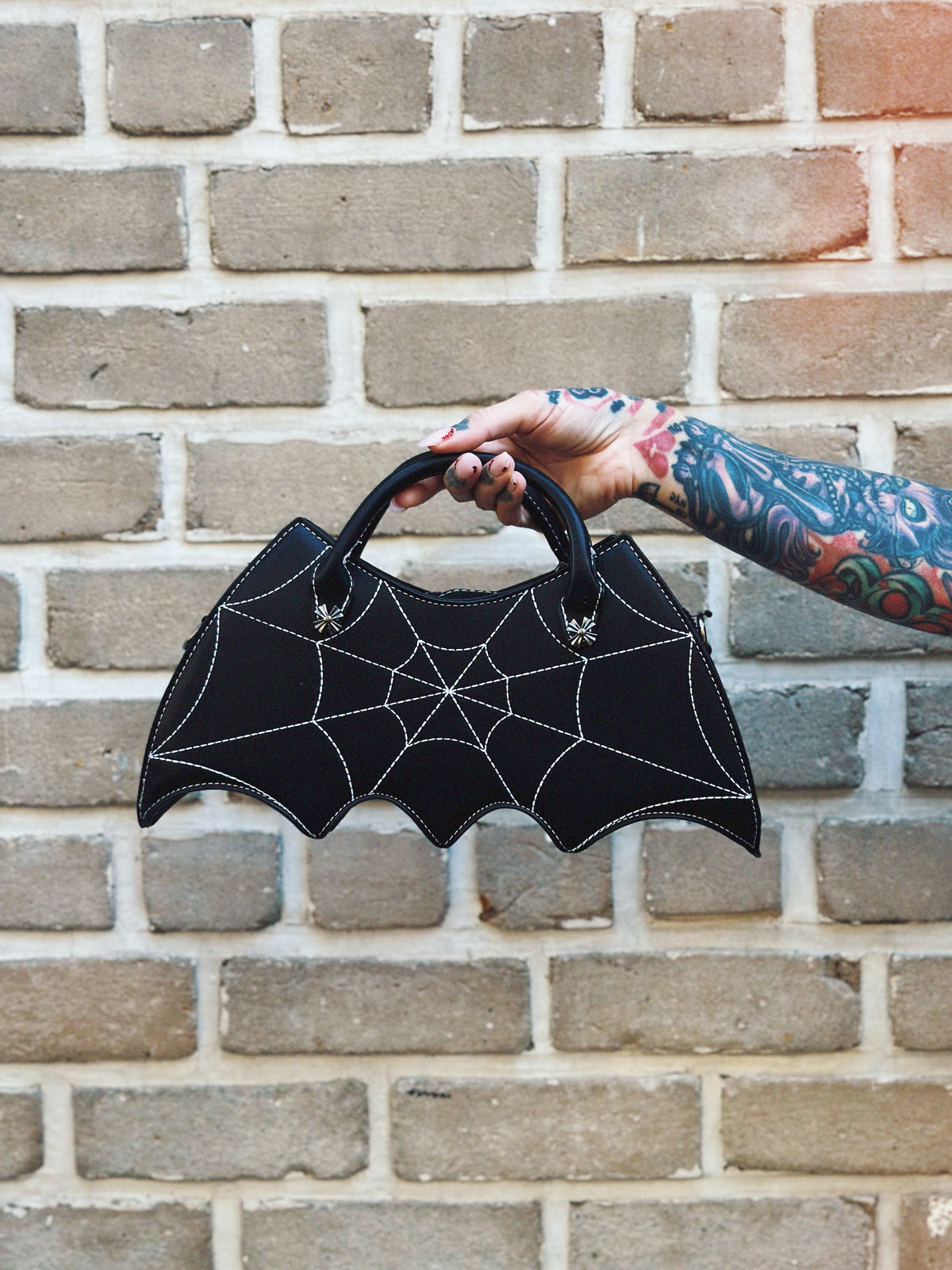 Black bat-shaped handbag with spider web design held by a tattooed arm against a brick wall.