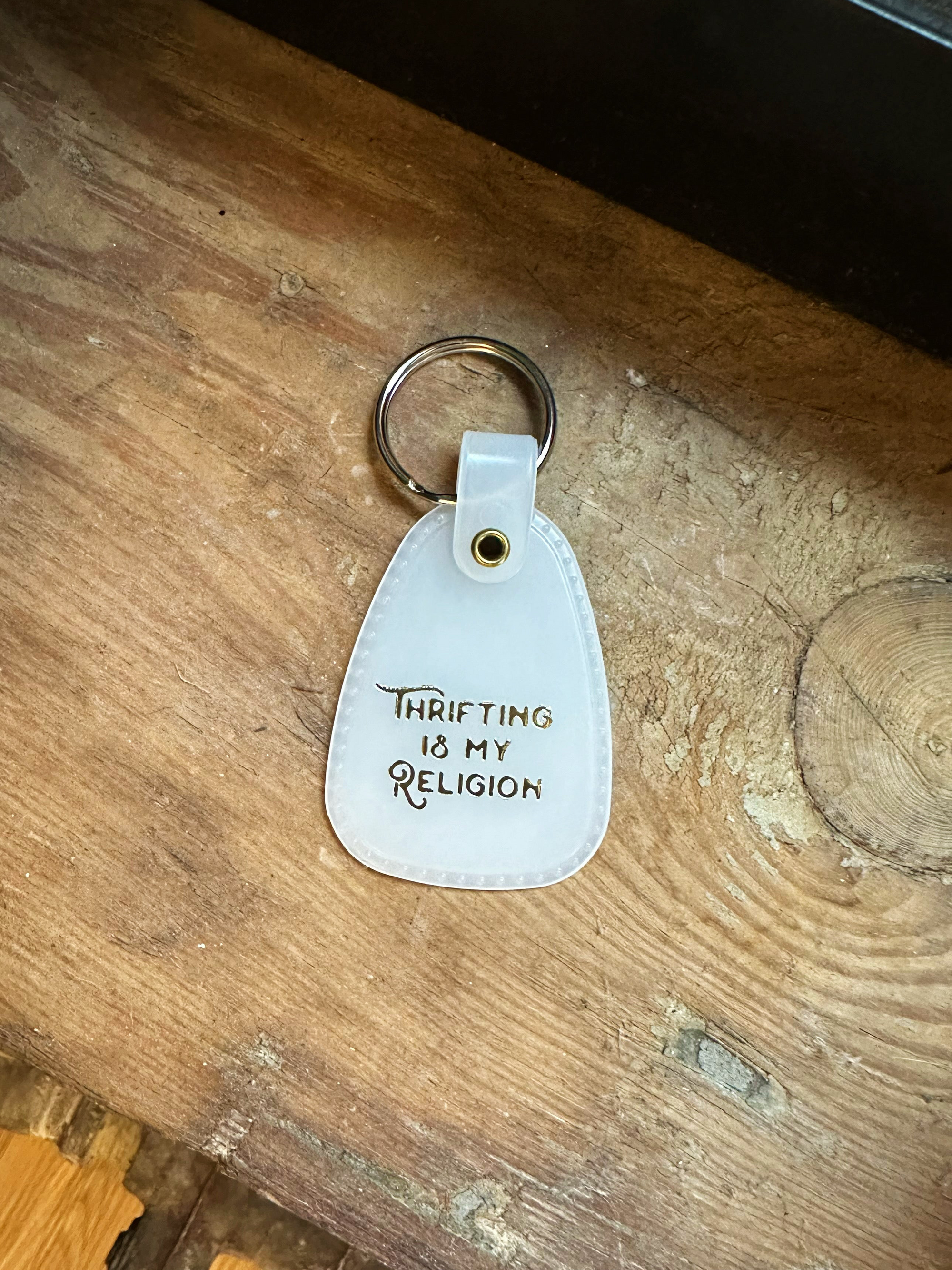 White saddle style keychain with 'Thrifting is my religion' text on a wooden surface