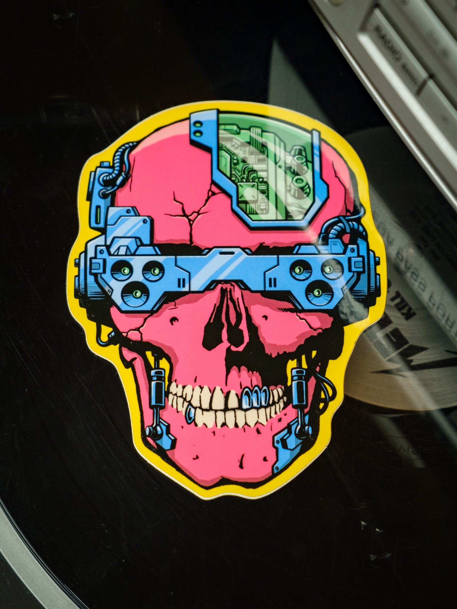 A colorful vinyl sticker depicting a skull with cyber punk aesthetics, including blue and yellow electronic components over a pink and red background.