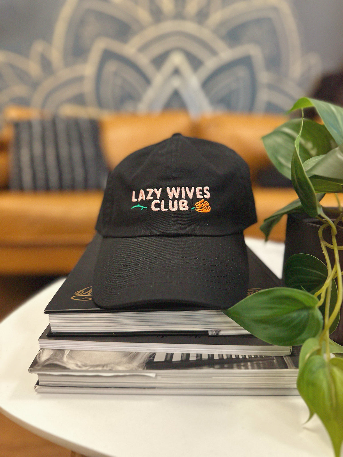 Black cap with 'Lazy Wives Club' text on a stack of books with a plant in the background