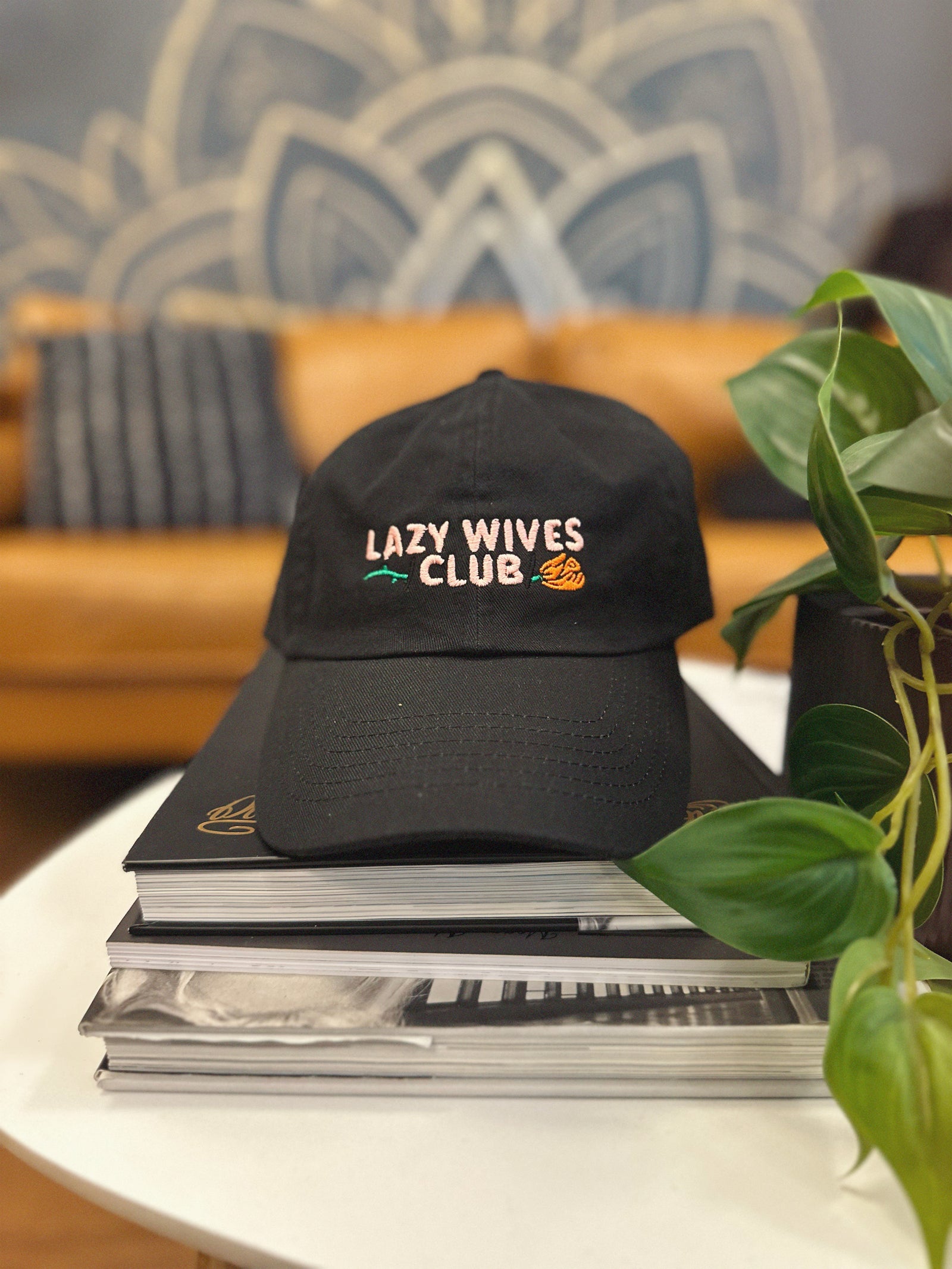 Black cap with 'Lazy Wives Club' text on a stack of books with a plant in the background