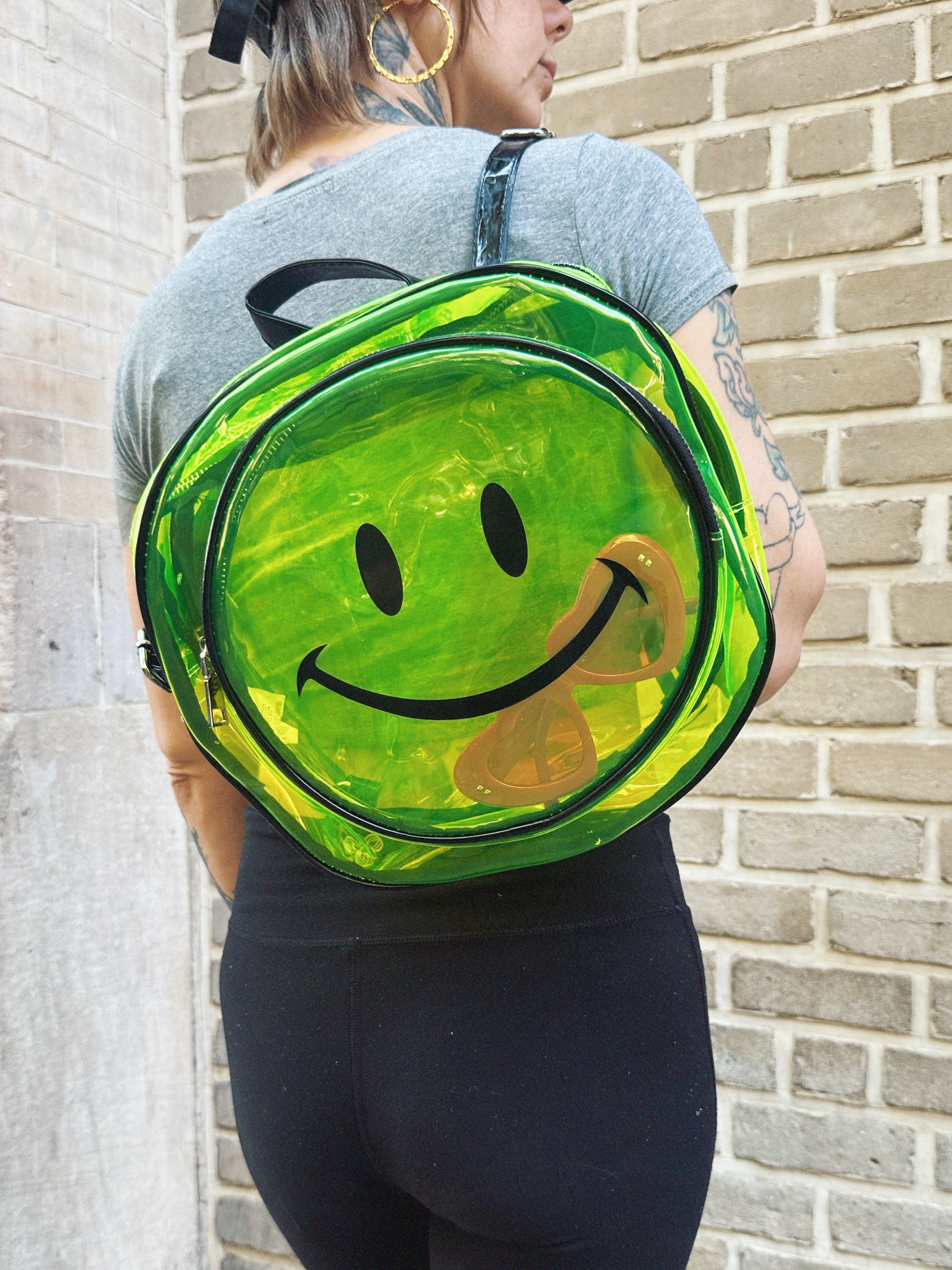 Person wearing a green smiley face backpack against a brick wall.