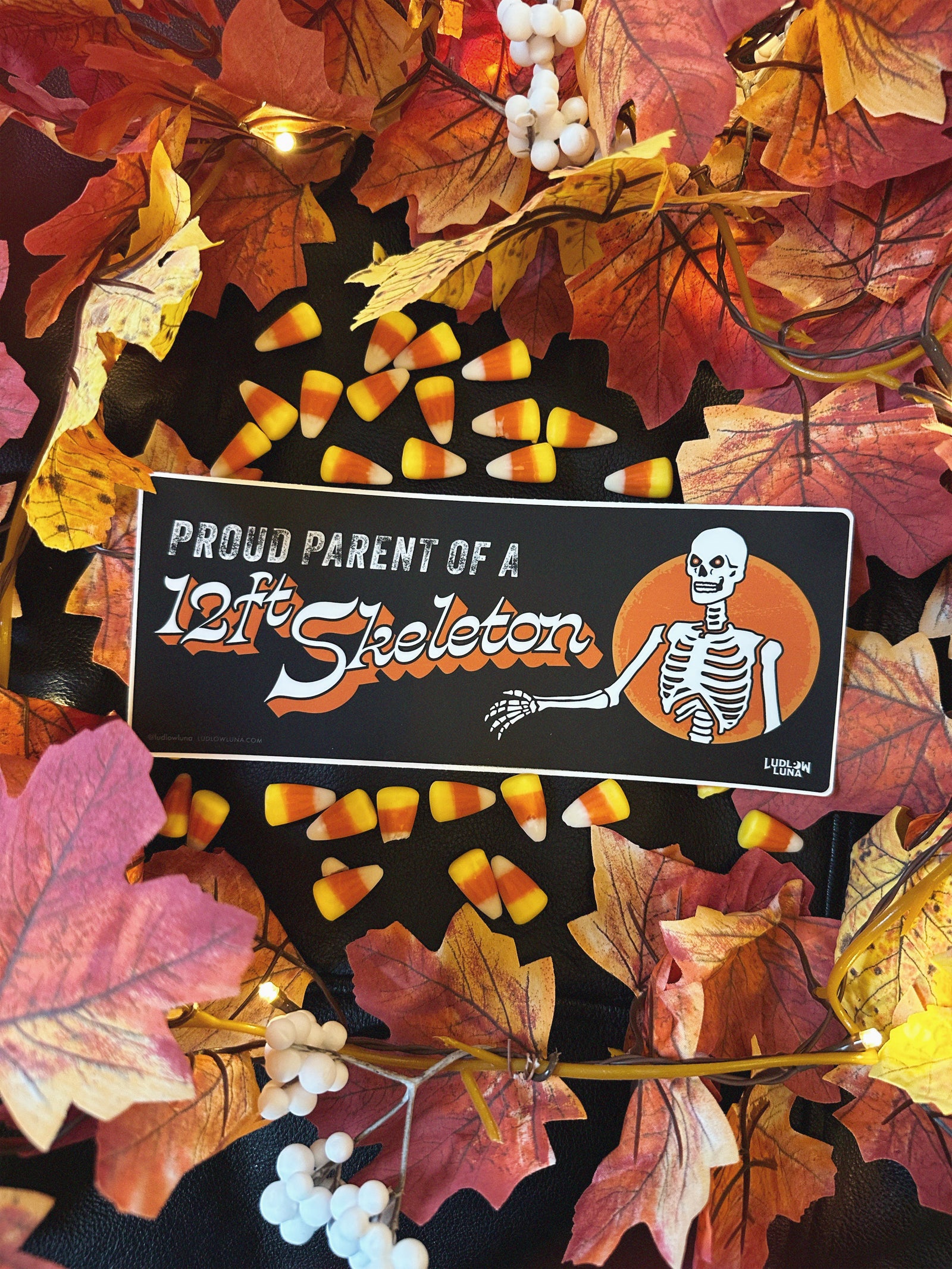 Halloween-themed orange bumper sticker with a skeleton graphic that reads "Proud Parent of a 12ft Skeleton" on a background with candy corn and autumn leaves.