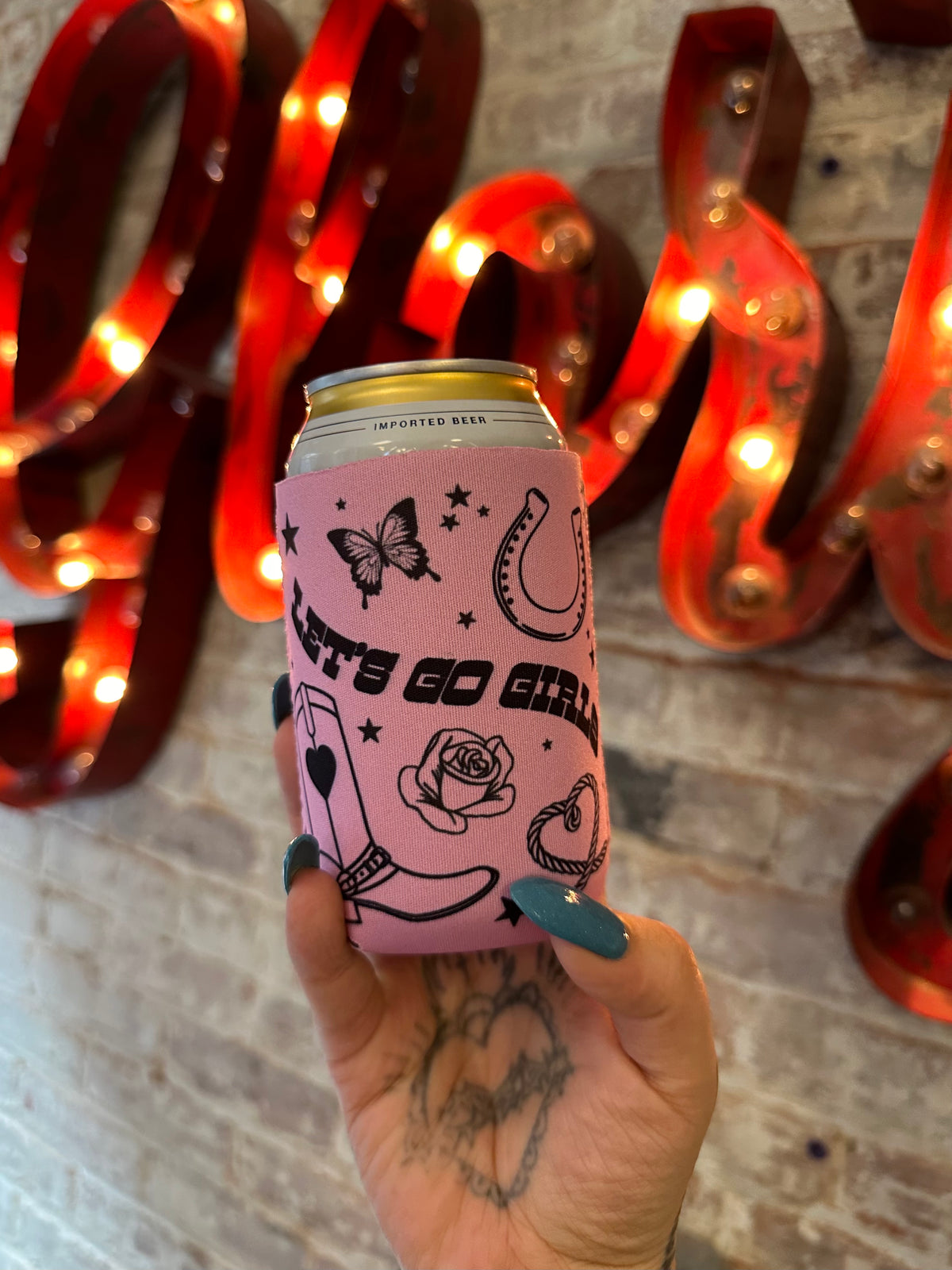 Pink can cooler with graphics held by a hand in front of decorative letters with lights.
