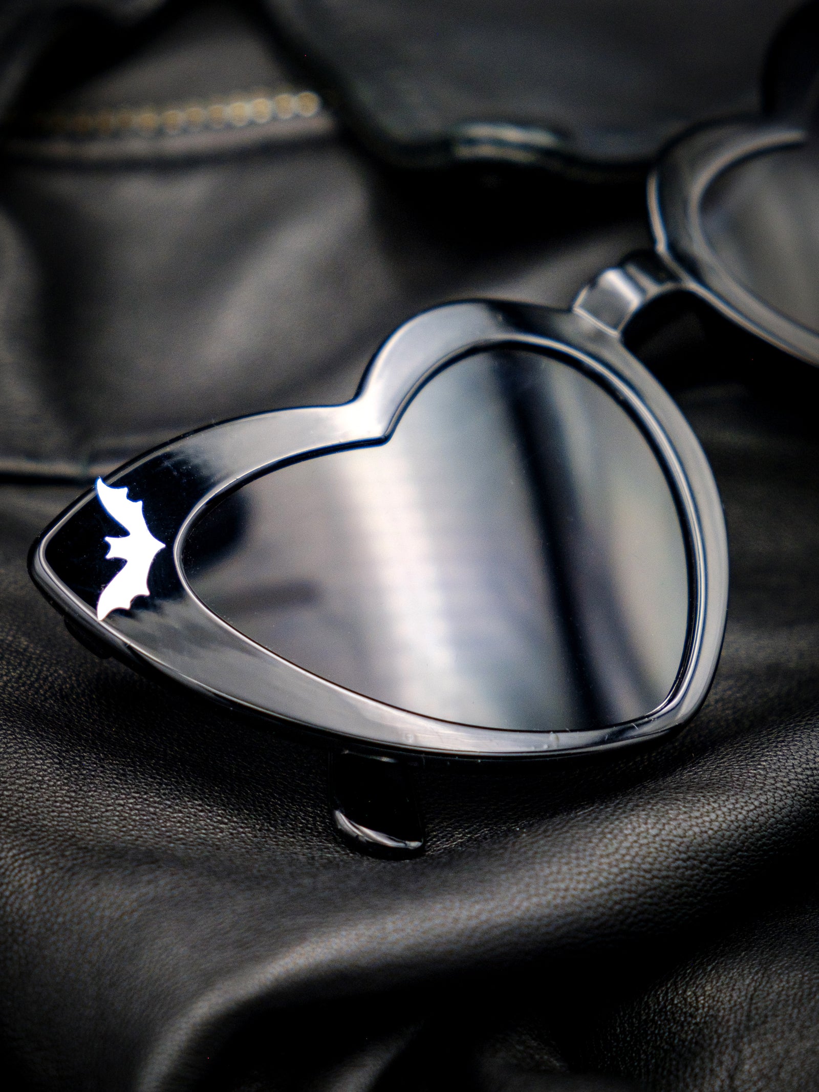Black heart-shaped sunglasses on a leather background. Sunglasses feature bat details in the corners and read Dead Inside along the arms. 