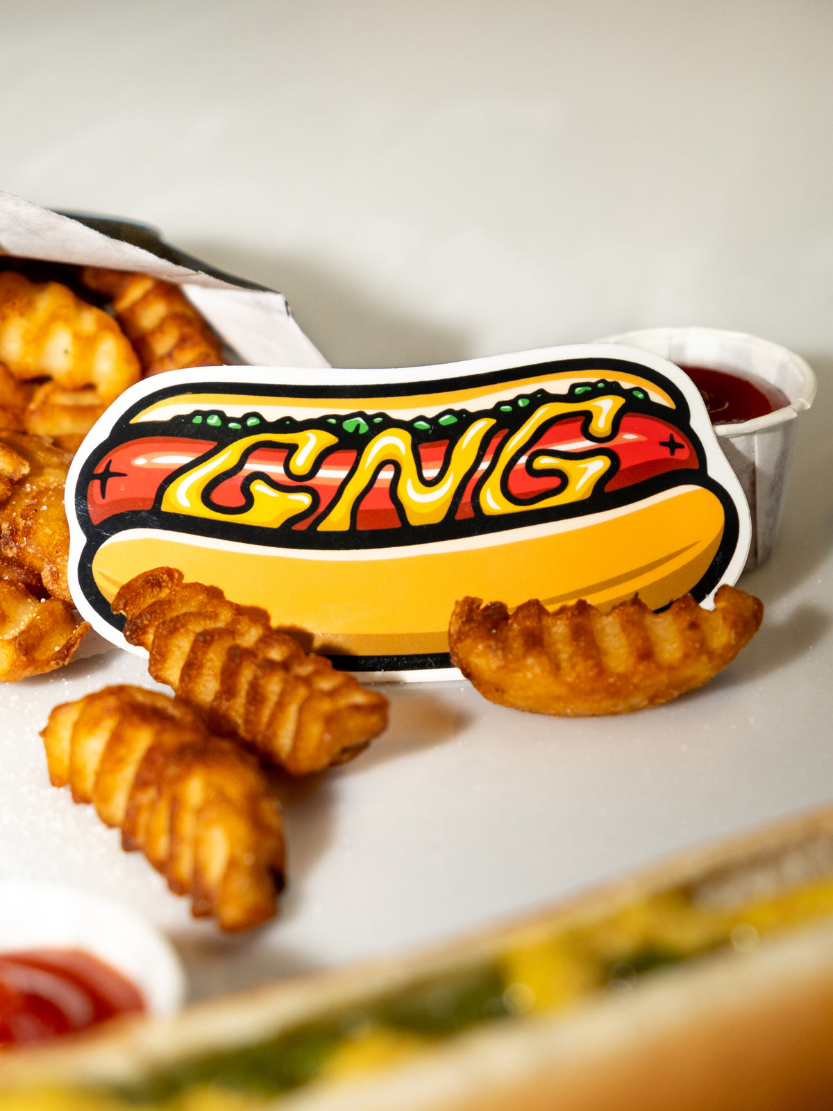A vinyl sticker with a hot dog design, labeled 'GNG', placed on a table with fries and ketchup.