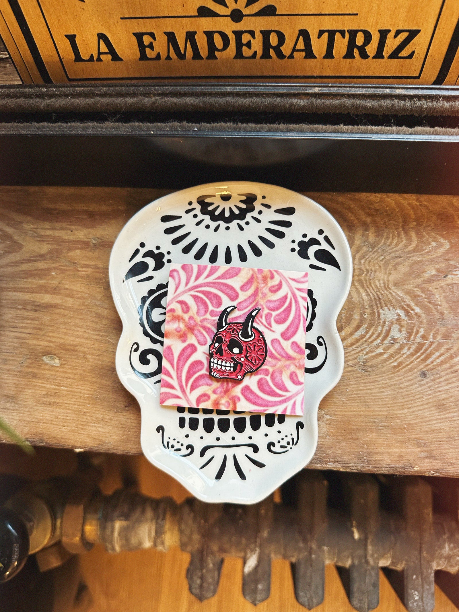 Decorative skull with pink and black design on a wooden surface with 'La Emperatriz' branding.