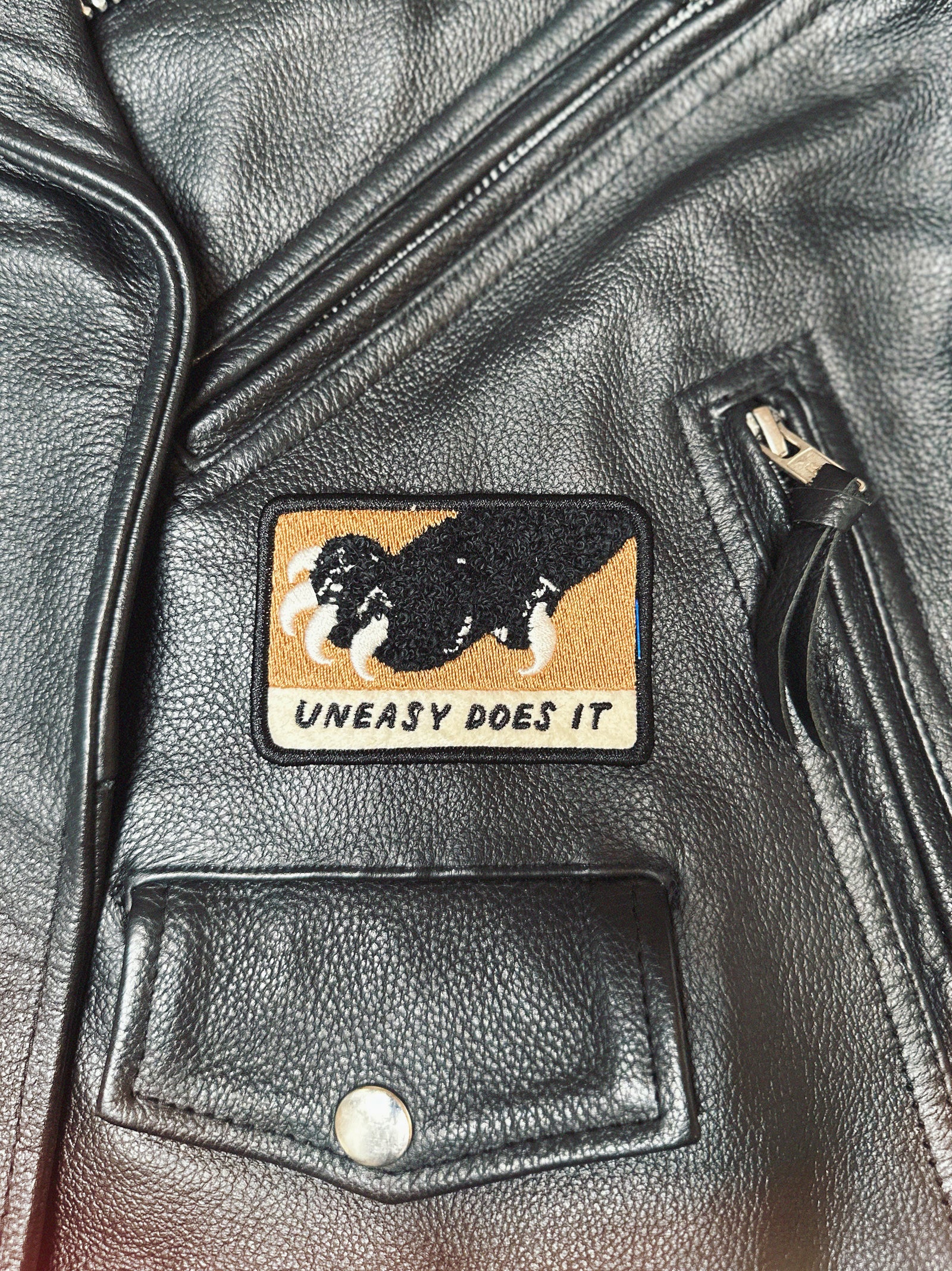 Black leather moto jacket with a patch featuring a paw with claws and text 'Uneasy Does It' on a brown background.