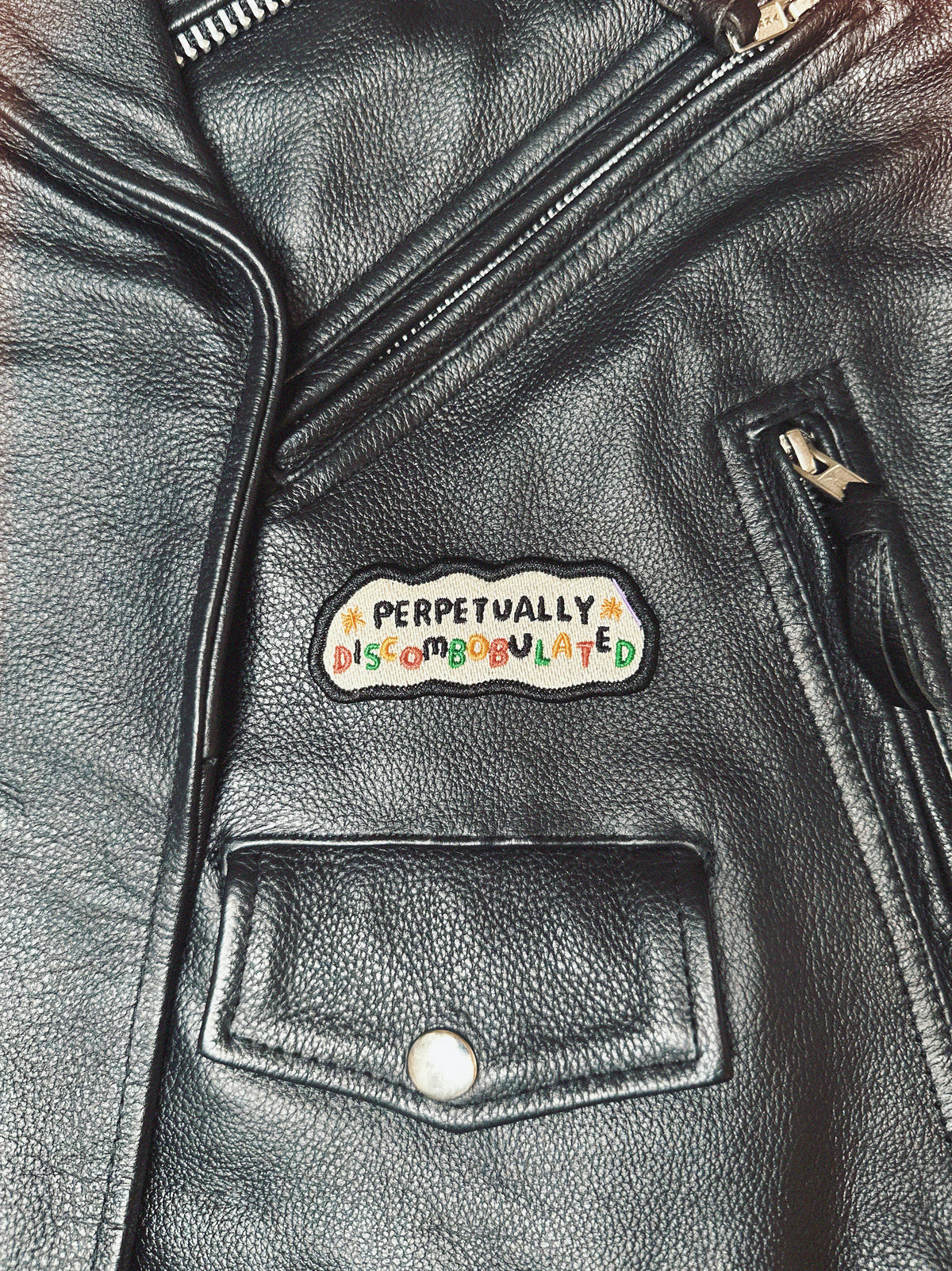 Black leather motorcycle jacket with a colorful patch on the chest that reads 'perpetually discombobulated'