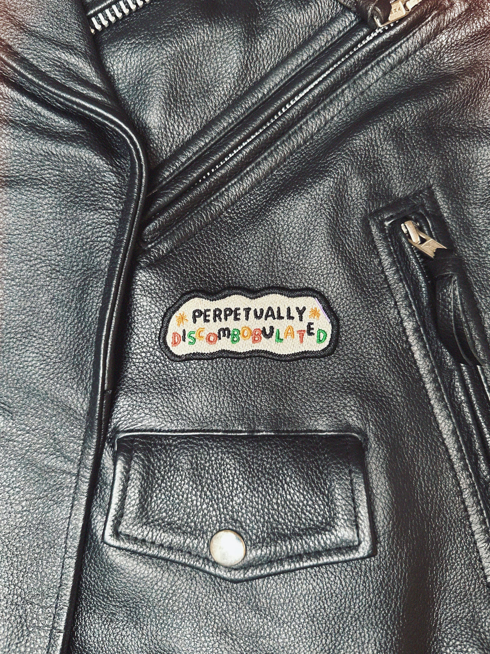 Black leather motorcycle jacket with a colorful patch on the chest that reads 'perpetually discombobulated'