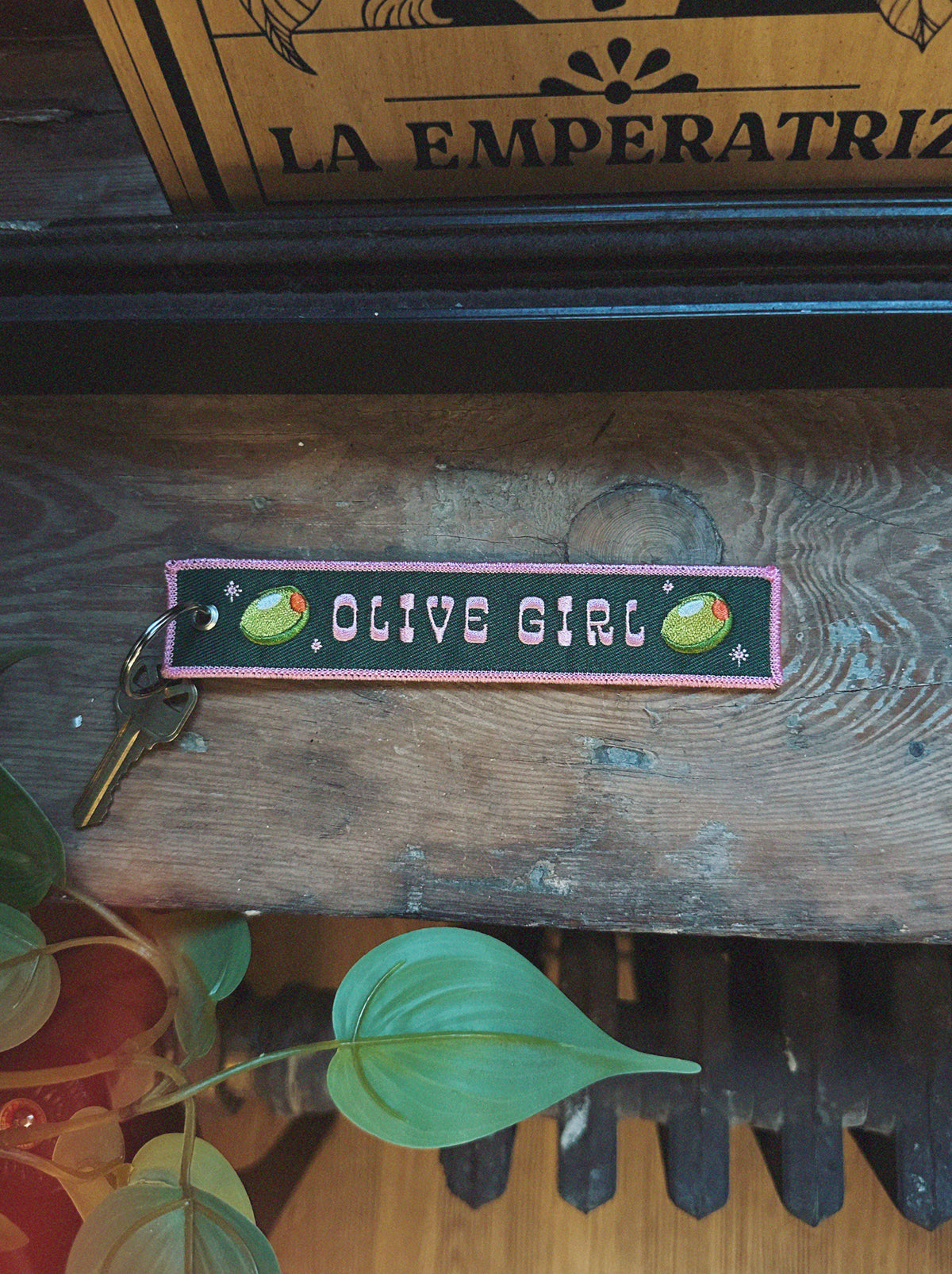 Embroidered keychain with 'OLIVE GIRL' text and olive graphics on a wooden surface