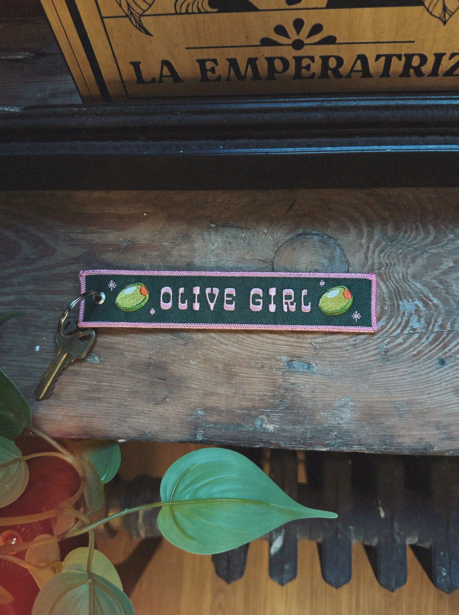 Embroidered keychain with 'OLIVE GIRL' text and olive graphics on a wooden surface