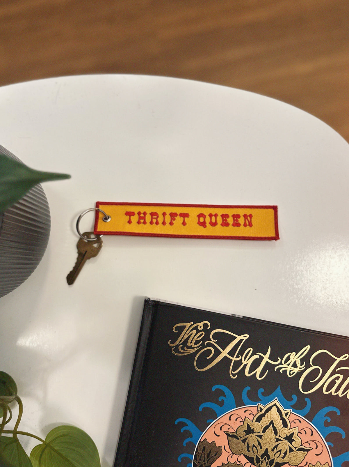 Yellow and red eychain with 'Thrift Queen' on a white surface with a book and plant