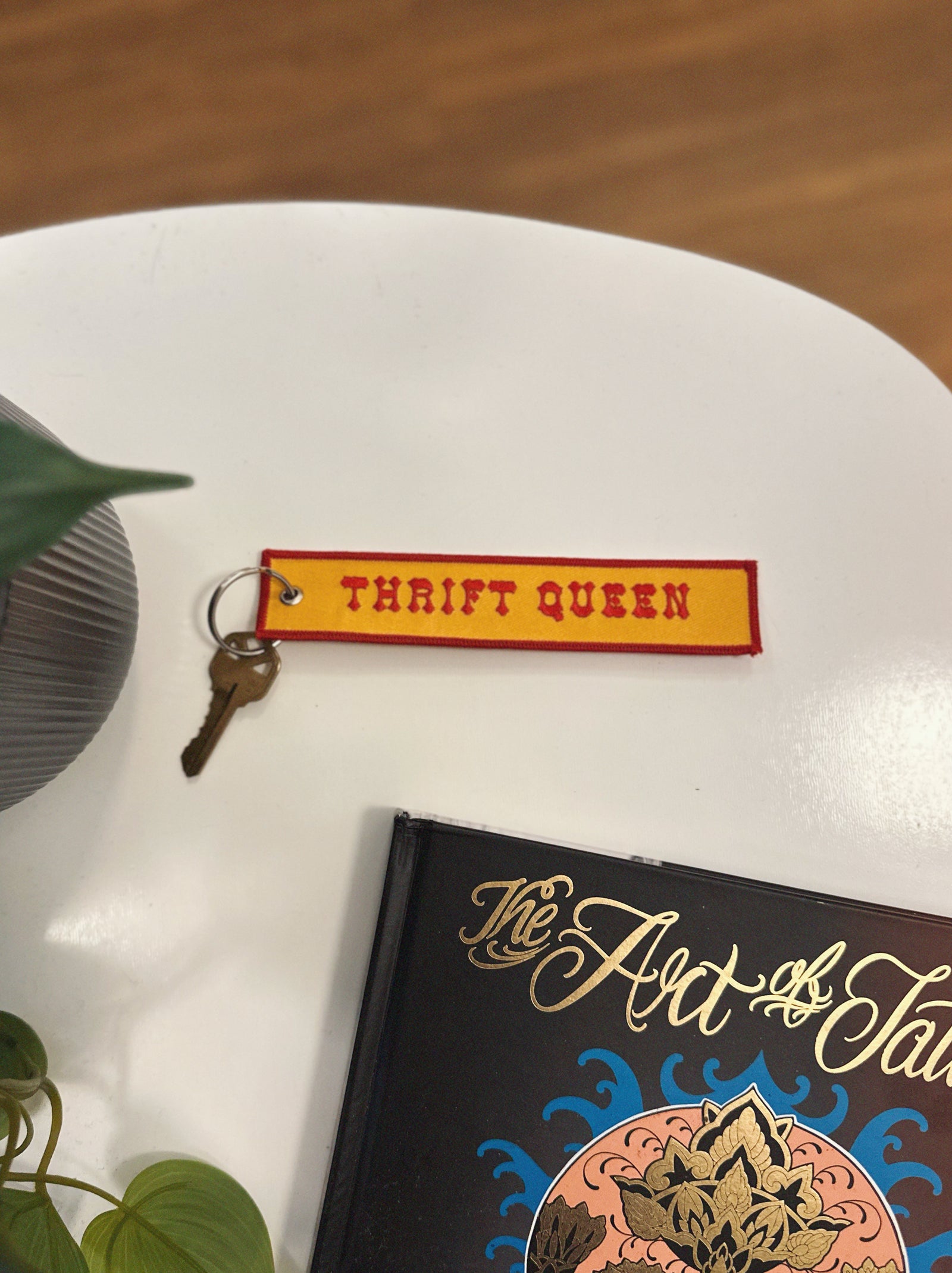 Yellow and red eychain with 'Thrift Queen' on a white surface with a book and plant